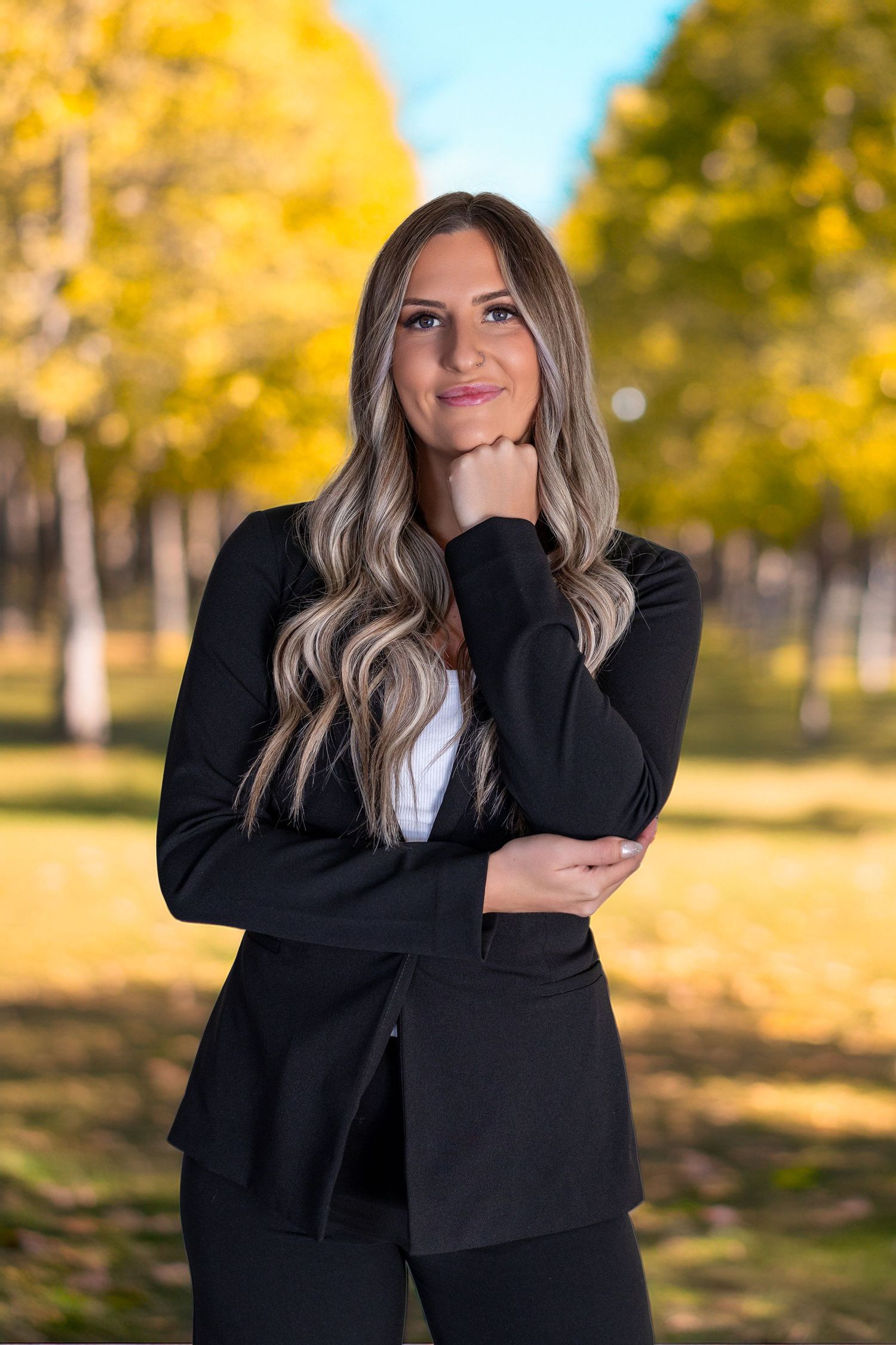 A professional woman in a black blazer poses thoughtfully in a park with vibrant yellow autumn trees in the background.