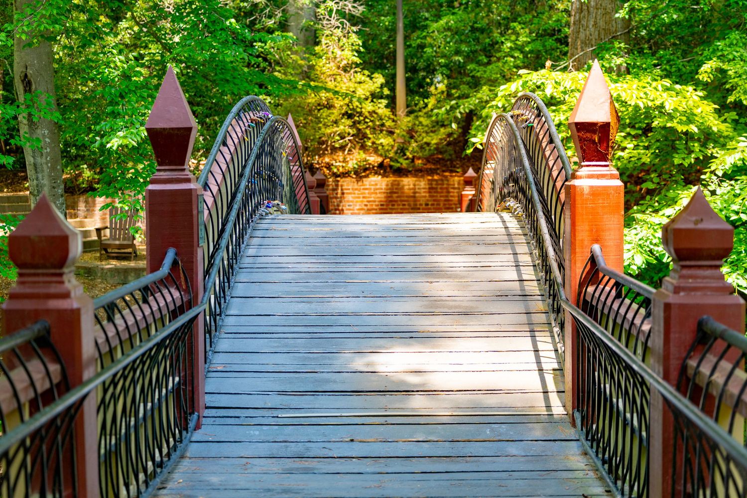 Wooden footbridge with ornate red iron railings leads through lush green forest park on sunny day.