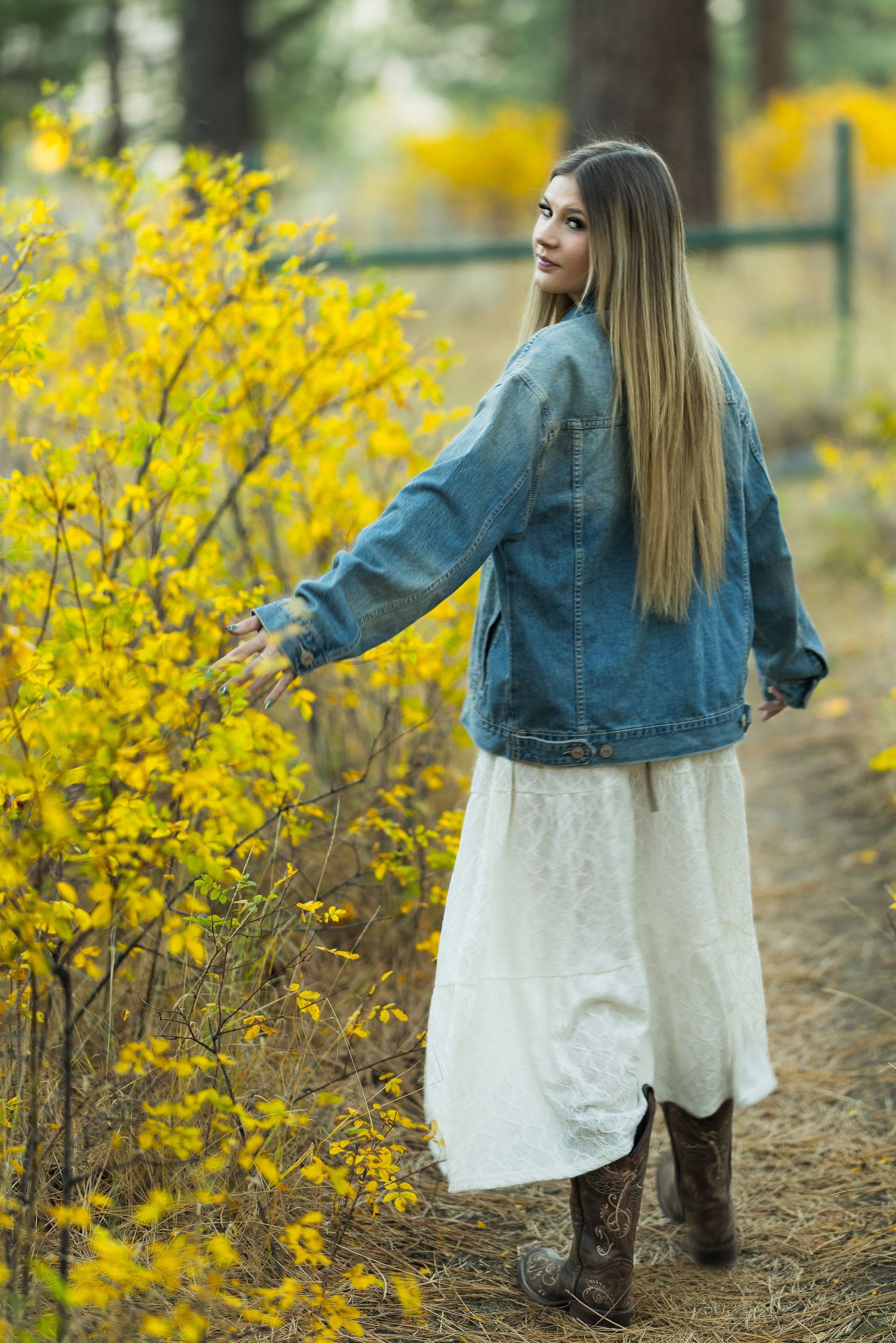 A person in a denim jacket and white dress stands on a forest path beside vibrant yellow forsythia bushes.