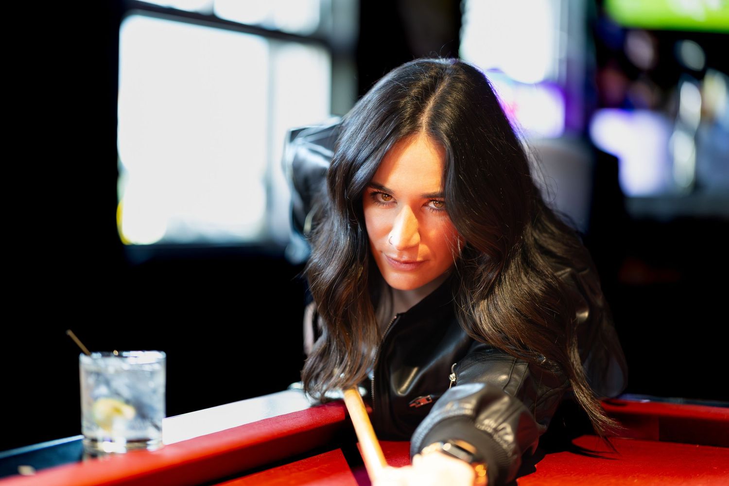 Woman with long dark hair leans over a red pool table, smiling while taking a shot in a dimly lit bar.