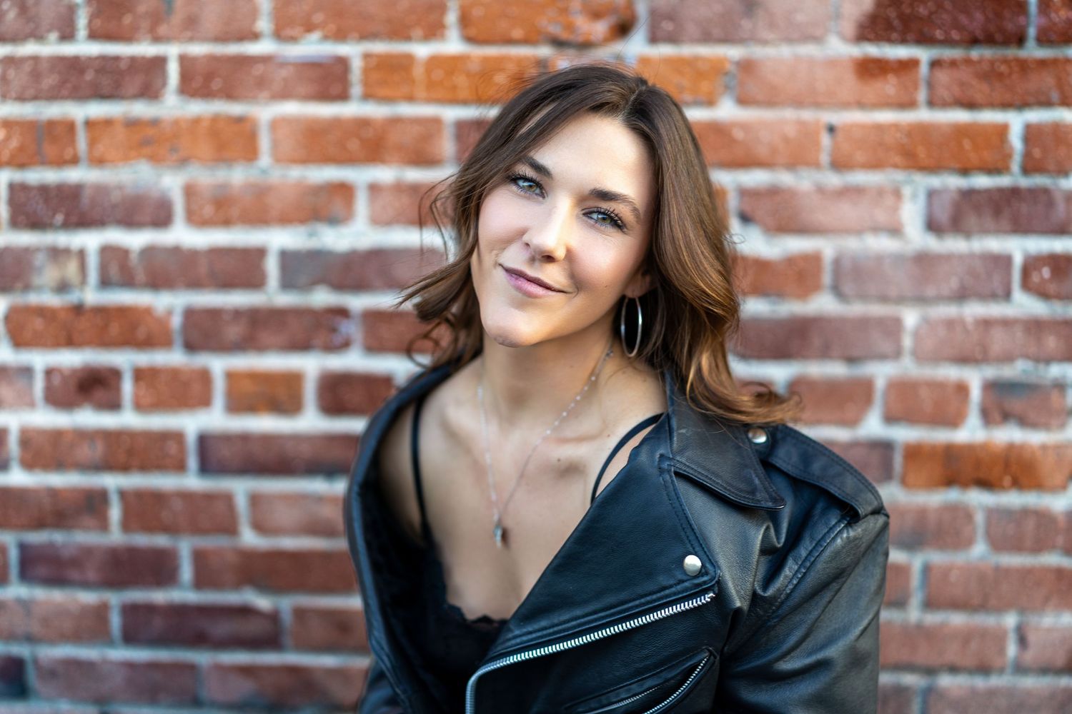 Person in black leather jacket poses against rustic brick wall in urban fashion photoshoot.