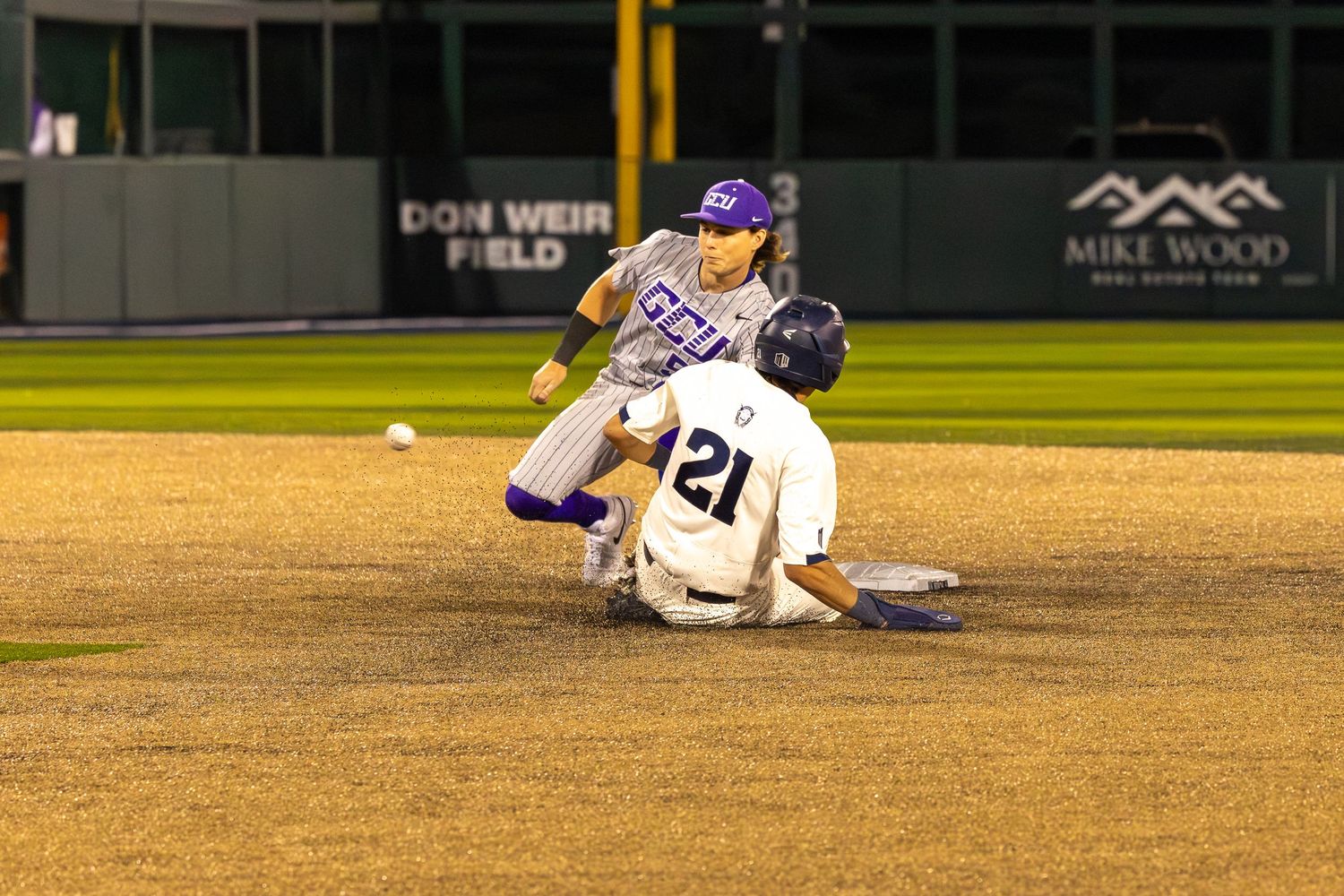 Baseball player sliding into second base as fielder attempts tag during night game at Don Weir Field.