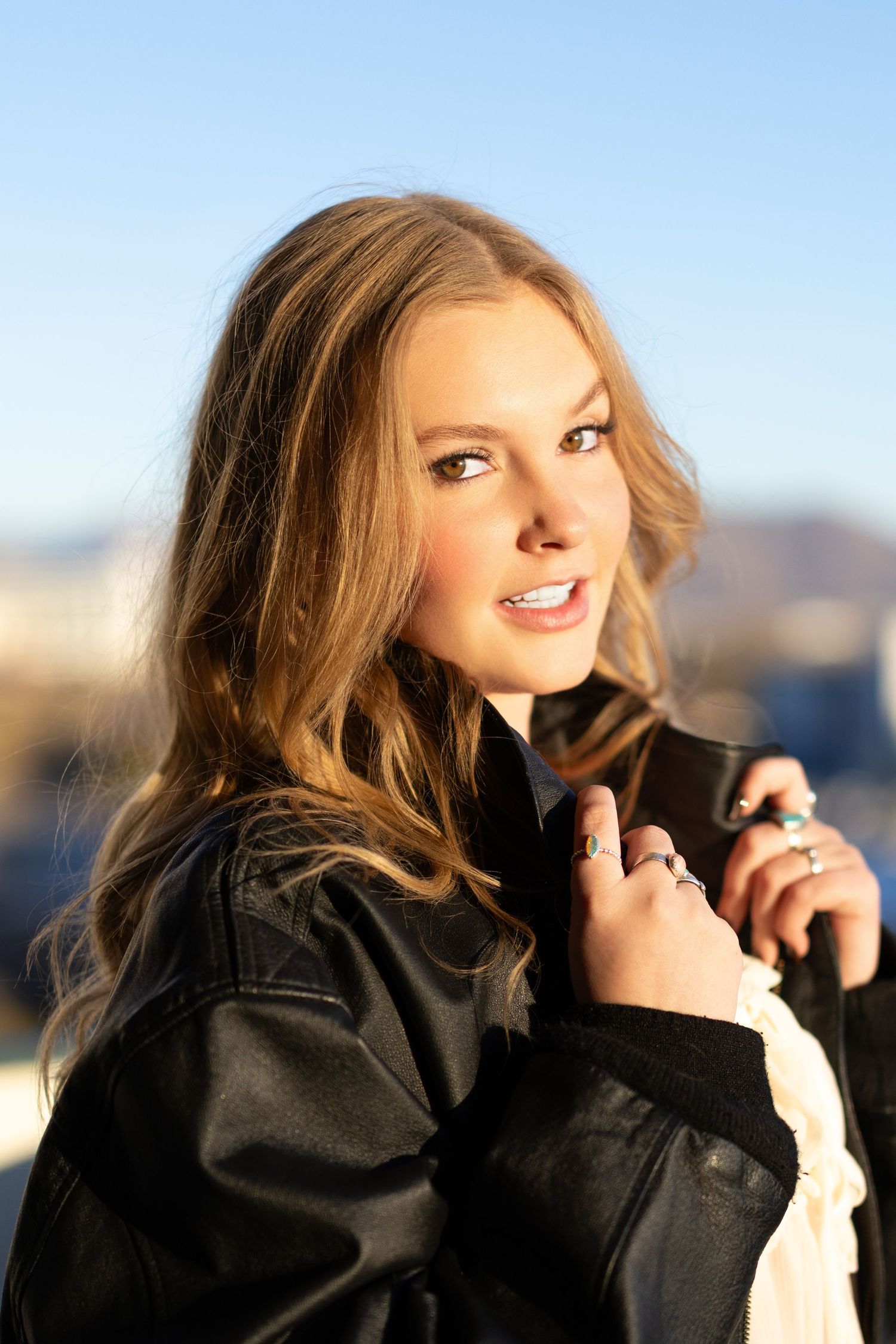Young woman with long blonde hair wearing a black leather jacket poses outdoors in bright sunlight.