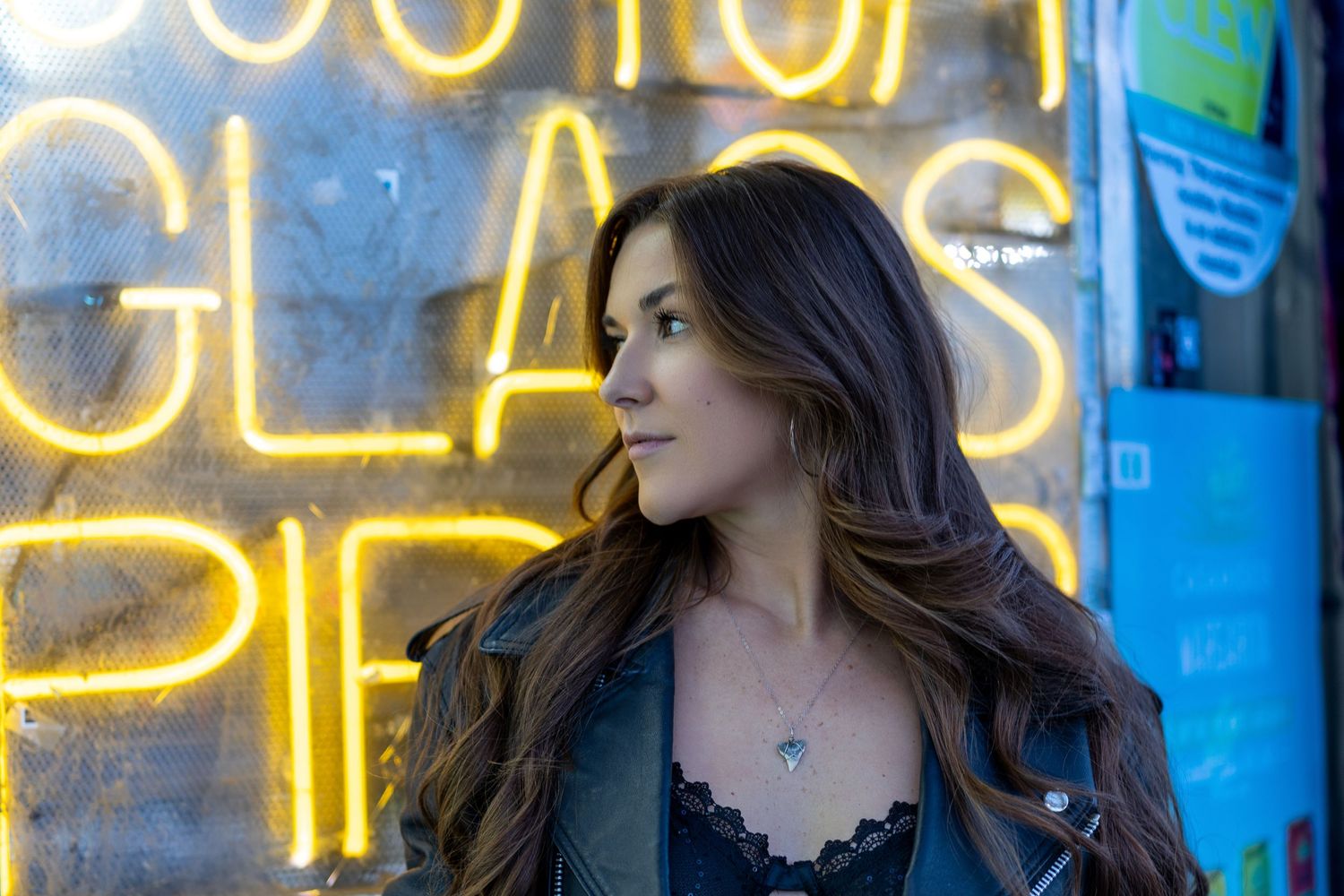 Young woman with long brunette hair poses in front of neon sign wearing leather jacket and delicate necklace.