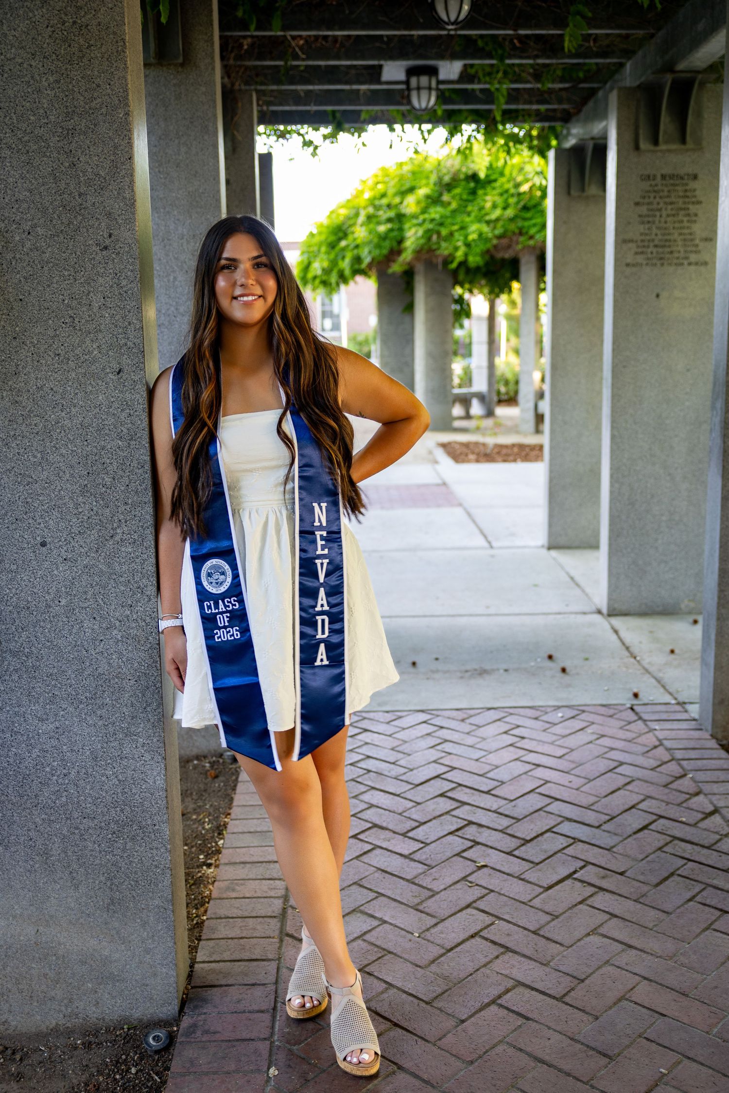 Young woman in white dress wearing a blue Nevada Class of 2025 graduation stole, posing outdoors on a brick walkway.