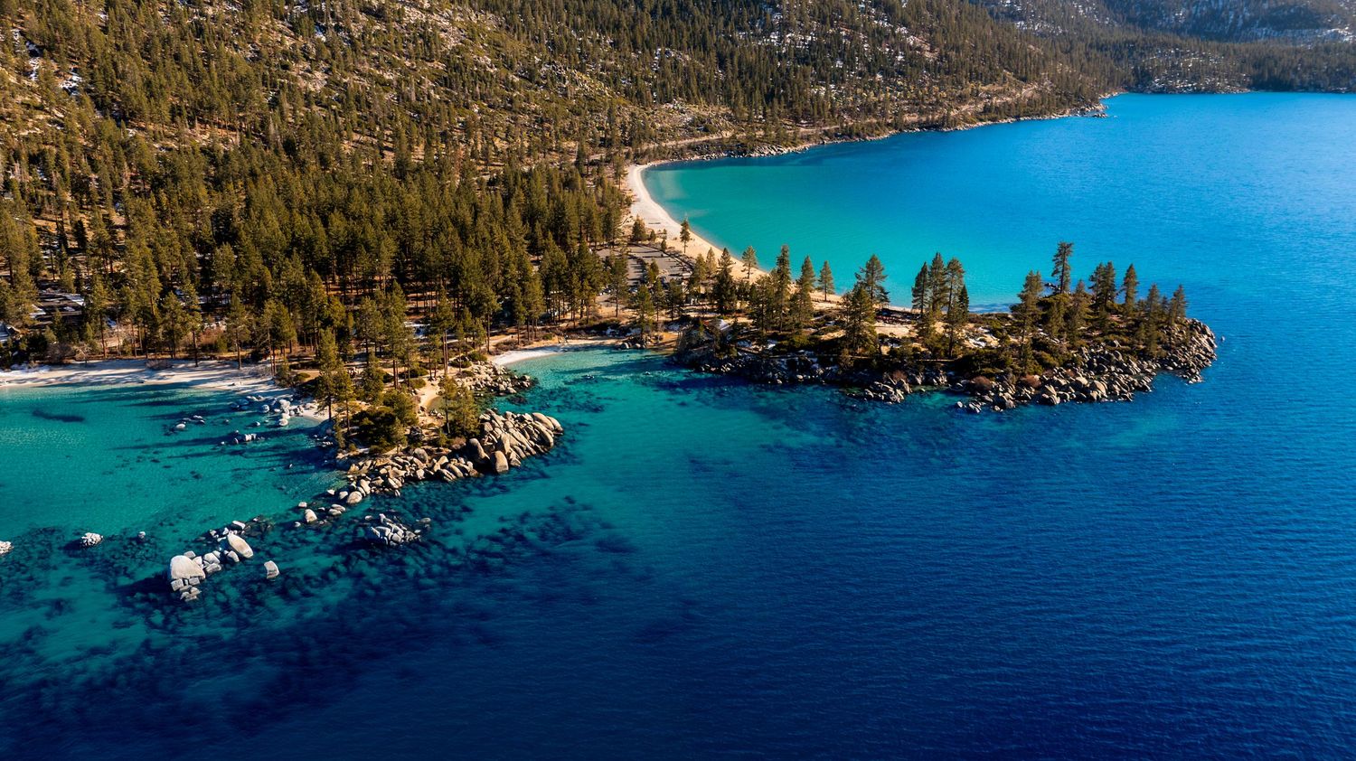 Aerial panoramic view of Lake Tahoe shoreline with turquoise waters, pine forests, and rocky outcrops on a sunny day.