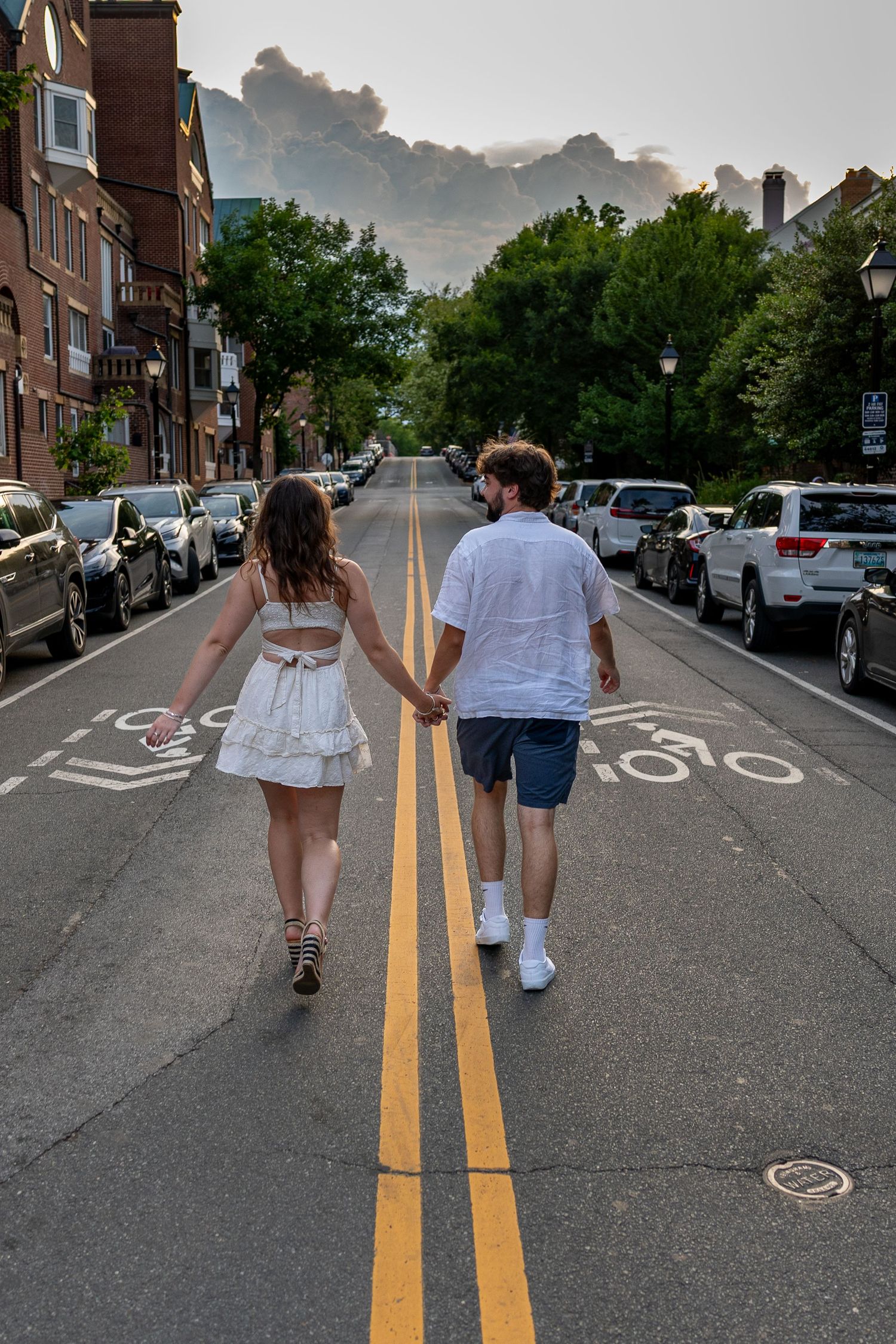 Couple holding hands walking down the middle of a tree-lined street in white outfits with mountains in the background.