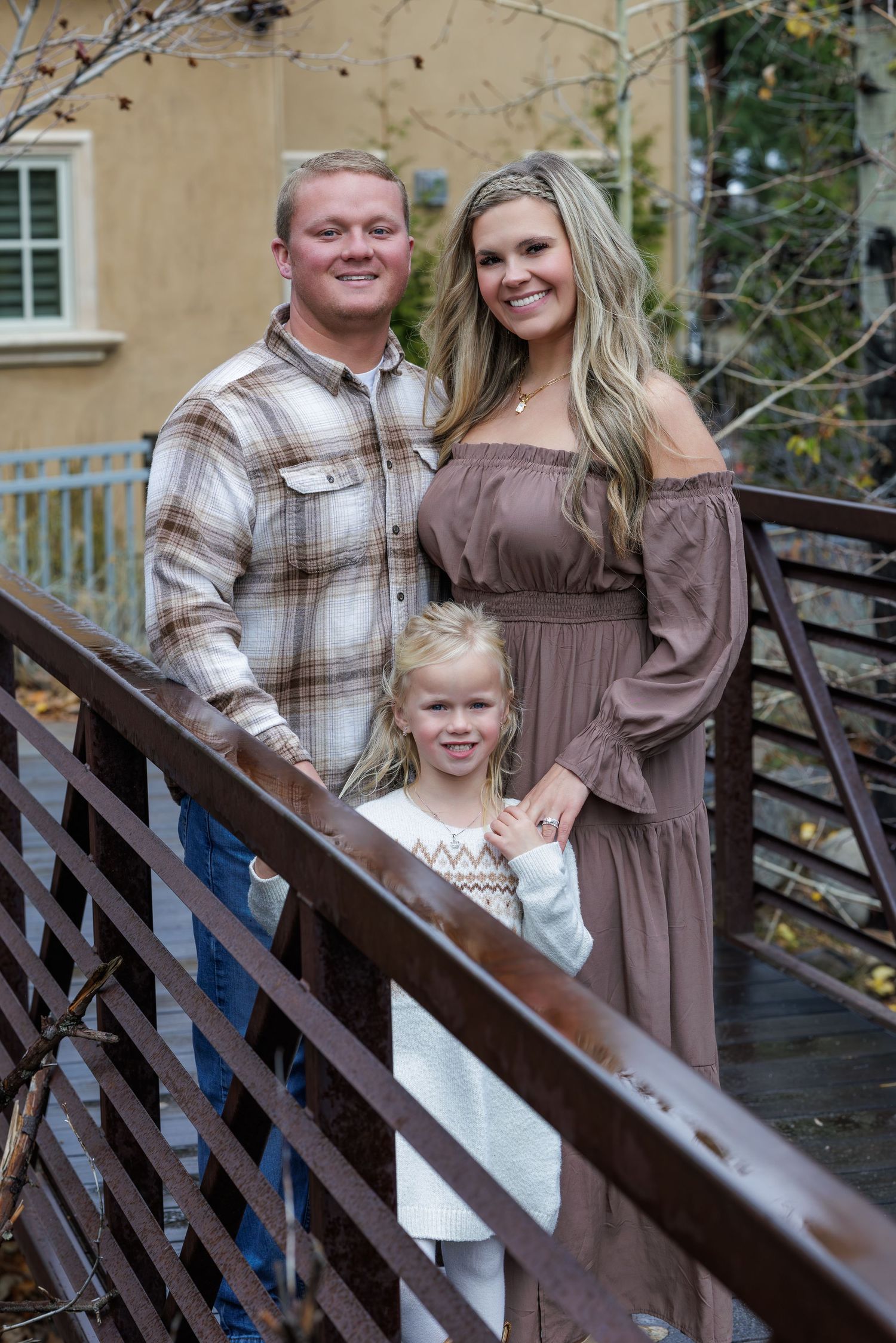 A family poses together on wooden stairs wearing coordinated brown and plaid outfits in an outdoor setting.