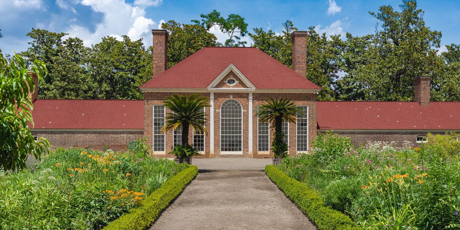 Historic red brick colonial building with palm trees, manicured hedges, and colorful garden path on a sunny day.