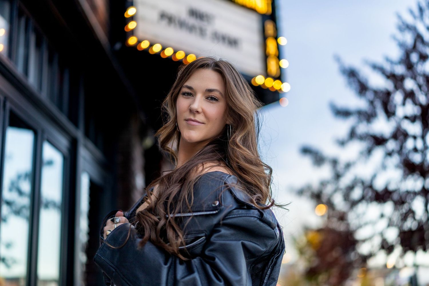 Stylish person in a black leather jacket poses on city street at dusk with illuminated storefronts in background.