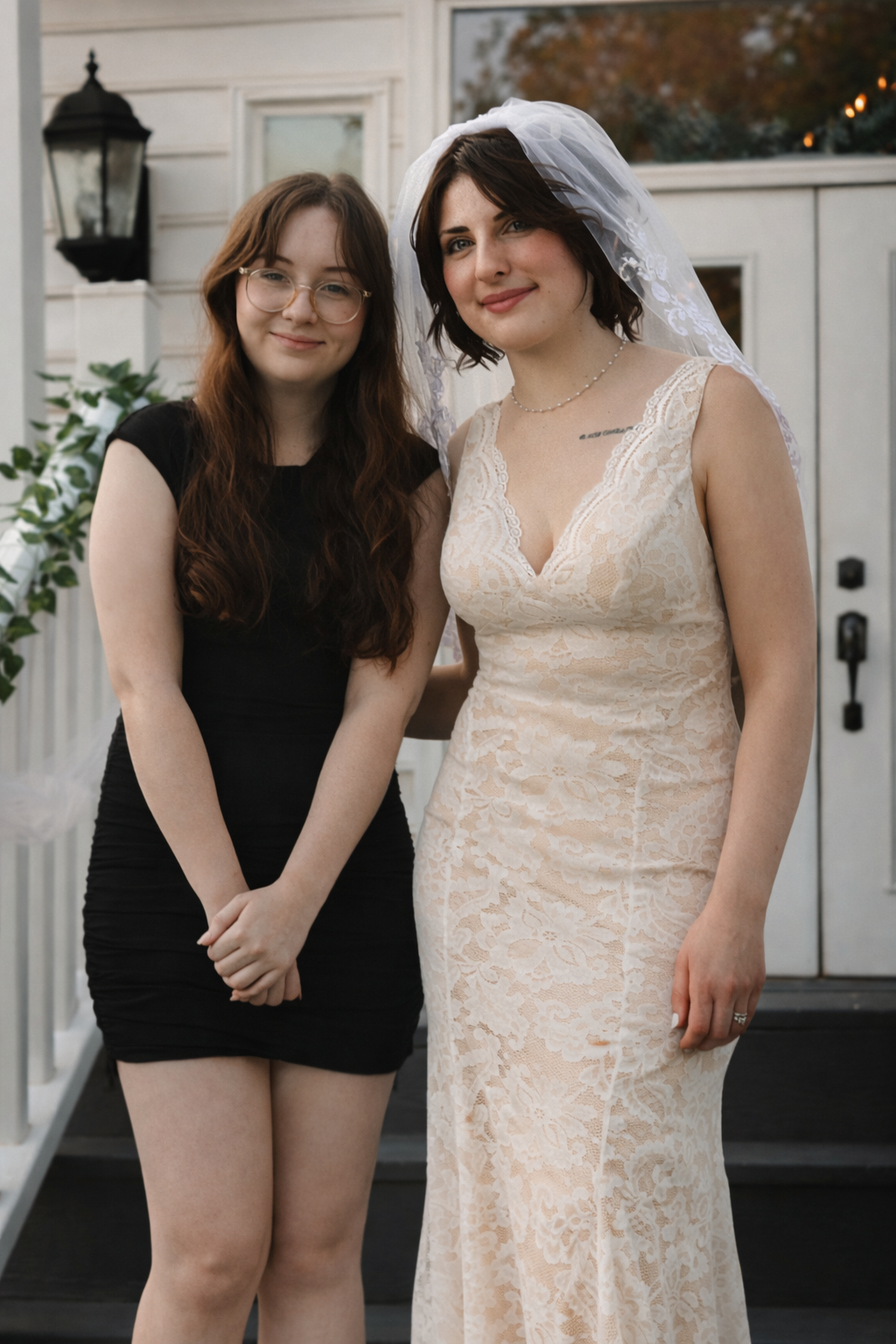 A bride in a cream lace dress poses with a companion in black on a white porch.