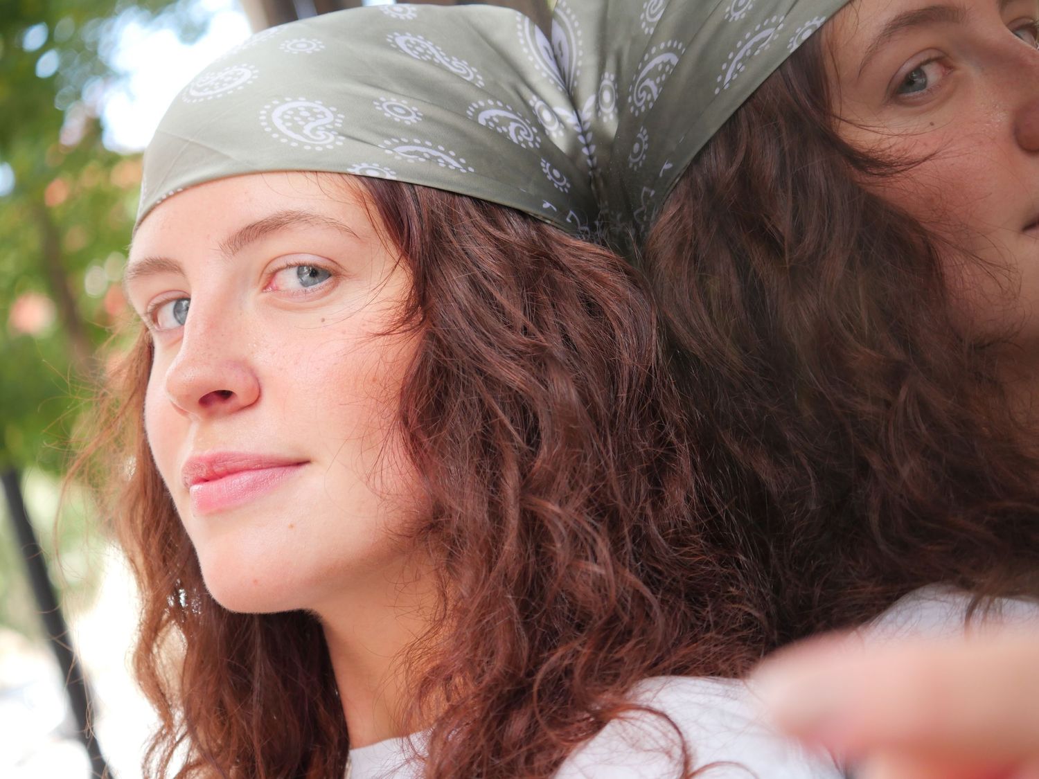 A woman wearing a light gray bandana with long wavy brown hair outside in natural lighting.