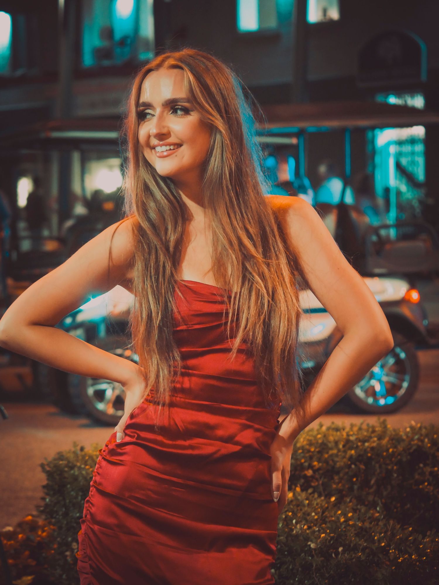 Woman in elegant red satin dress poses outdoors at night with urban lighting creating a moody atmosphere.