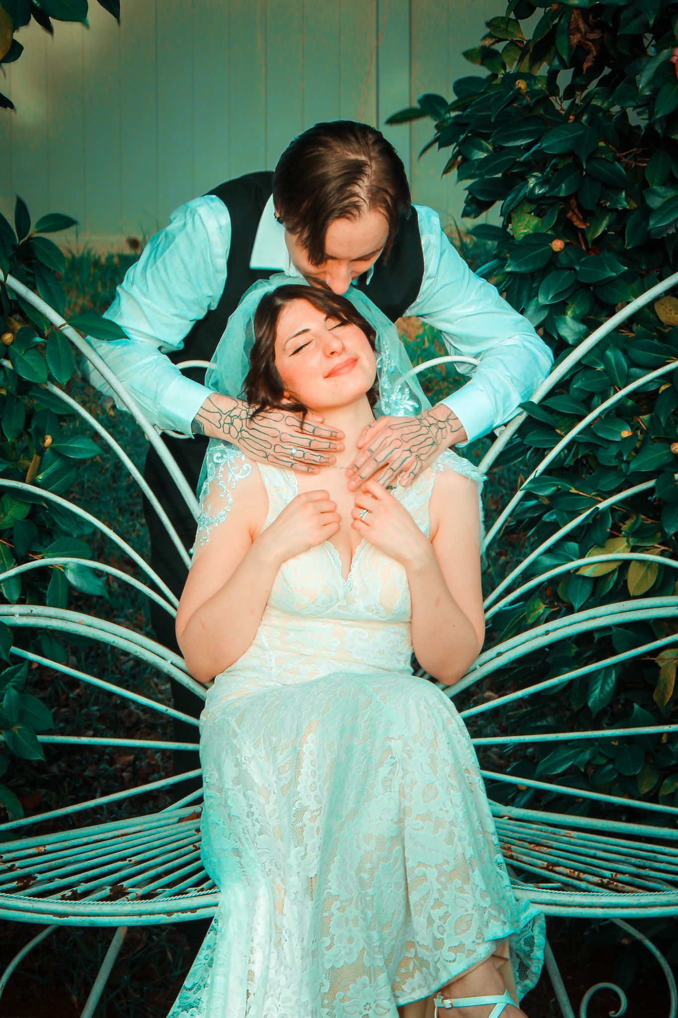 Couple in formal attire posing on decorative garden bench with turquoise lighting.