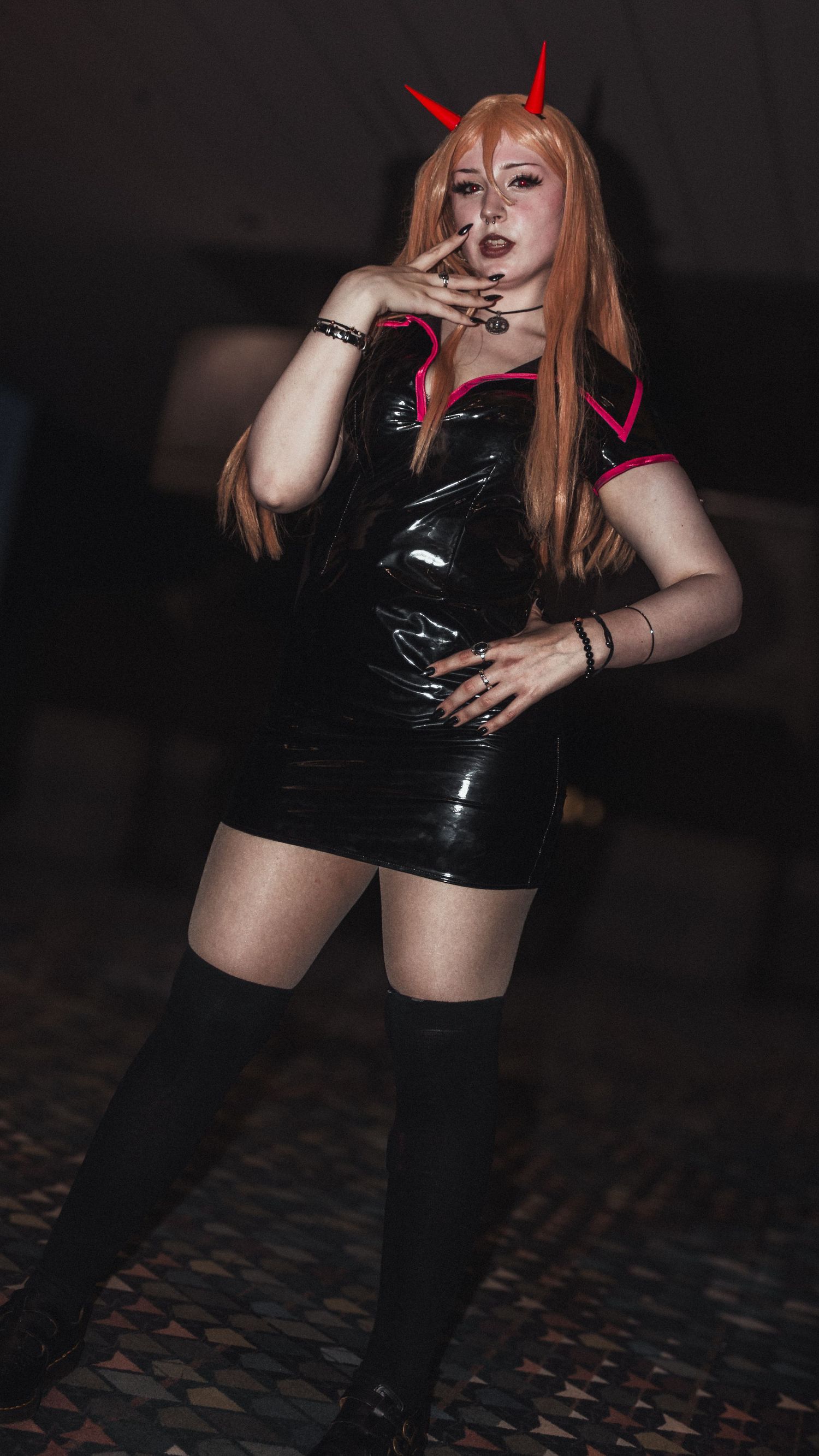Cosplayer in black vinyl dress and red horns strikes dramatic pose against dark background.