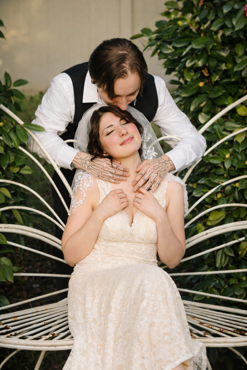 Person in a white lace dress poses elegantly on a white metal garden bench with greenery in the background.