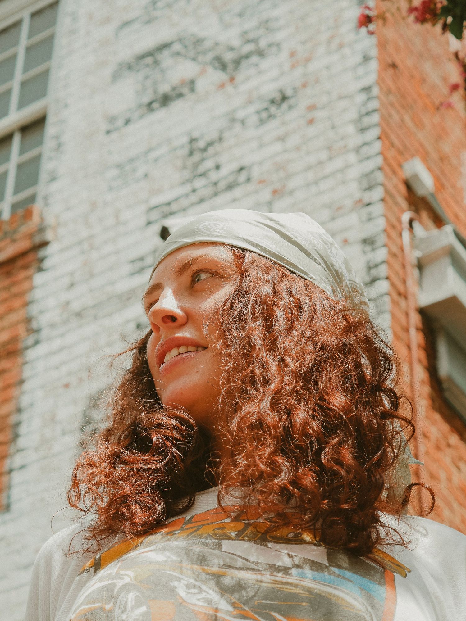 A person with curly auburn hair wearing a white beanie and top looks up at the sky against a brick building background.