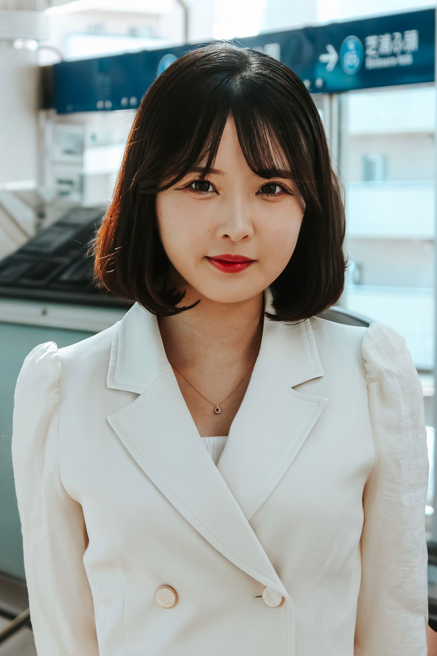 Professional portrait of someone in a white blazer with a necklace against a business interior background.