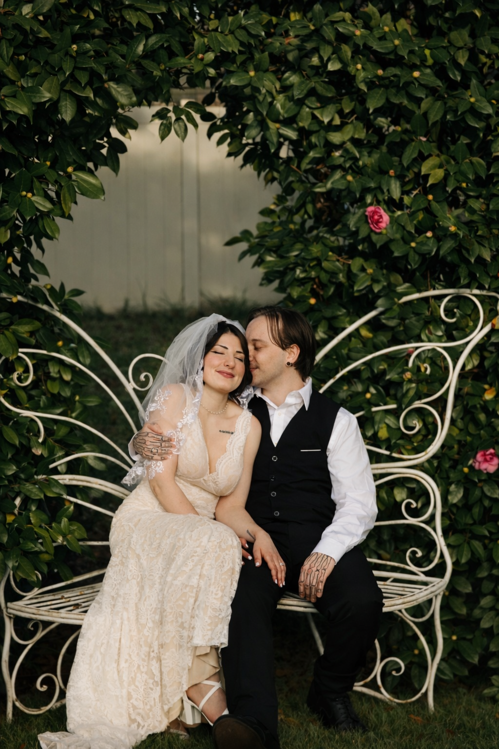 A romantic couple sits on a decorative white metal butterfly bench surrounded by lush greenery and pink roses.