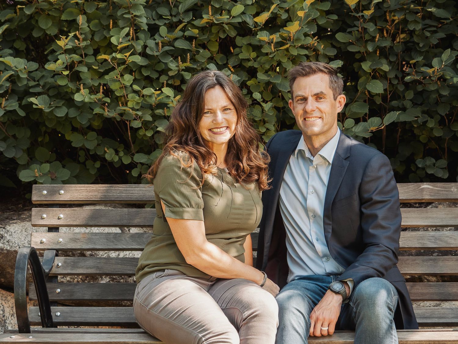 A couple sitting together on a wooden park bench with green foliage in the background.