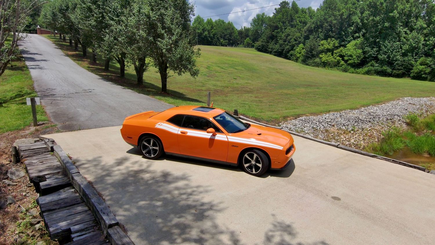 A panoramic view of an orange sports car parked on a curved driveway through wooded grounds.