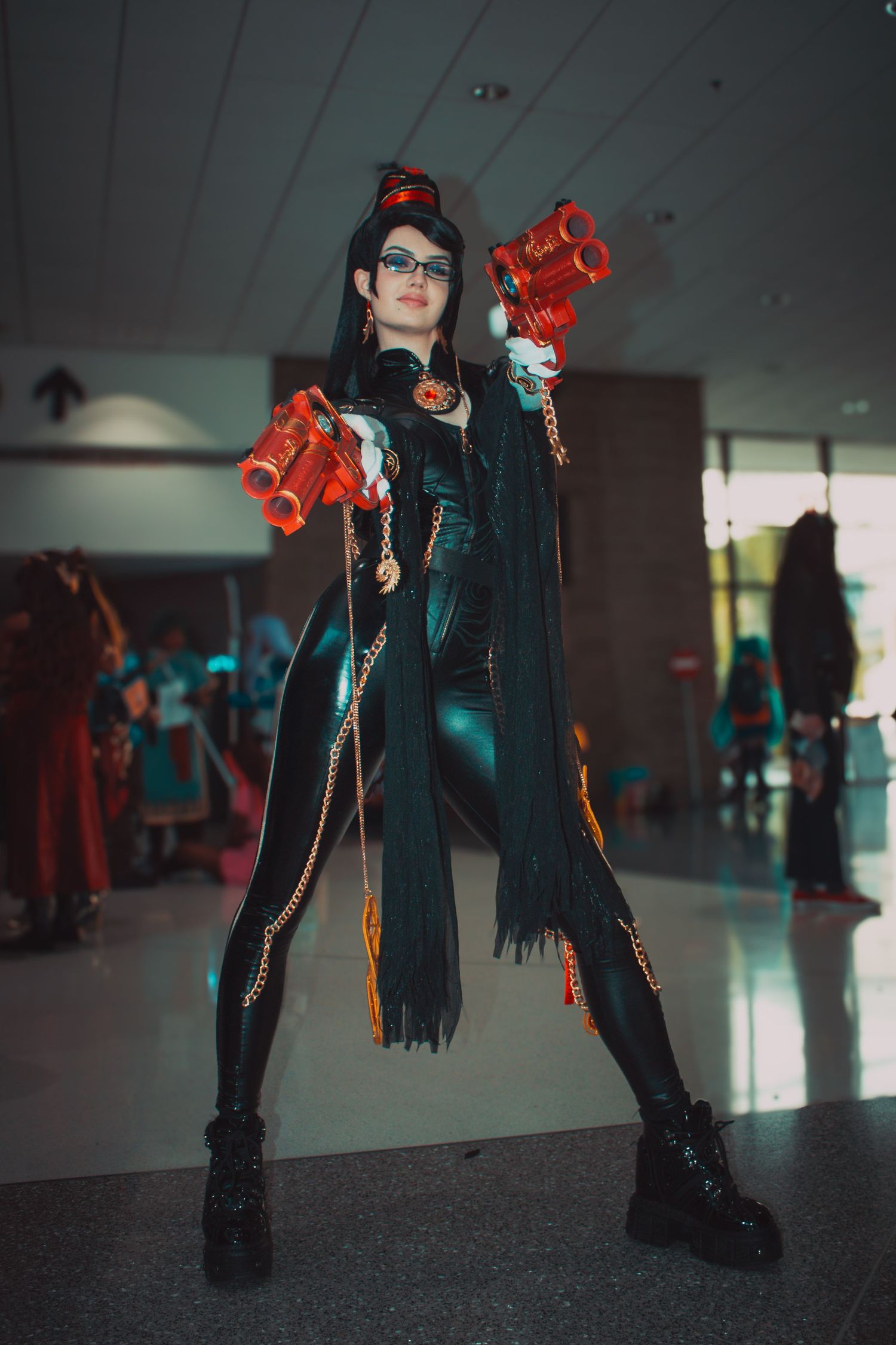 Figure in black leather costume poses with red prop weapons and chains in convention center hallway.