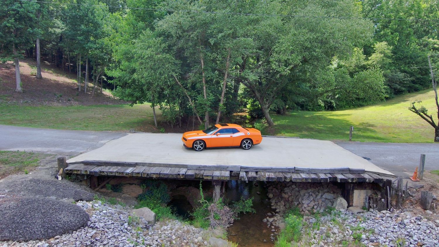 A bright orange classic muscle car on a winding driveway surrounded by lush green trees and landscaping.