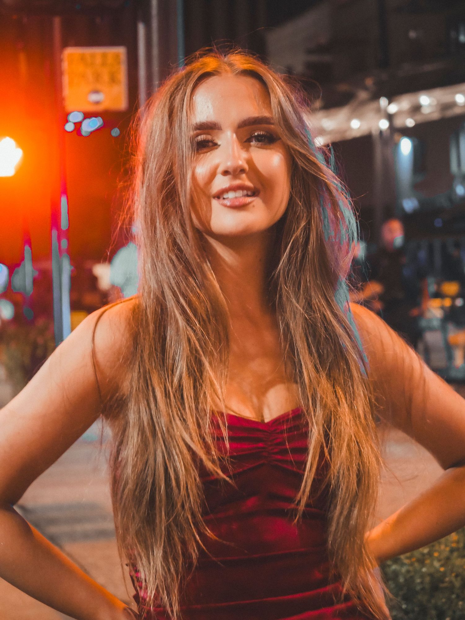 Woman with long brown hair wearing a red dress smiles under warm nighttime lighting in an urban setting.