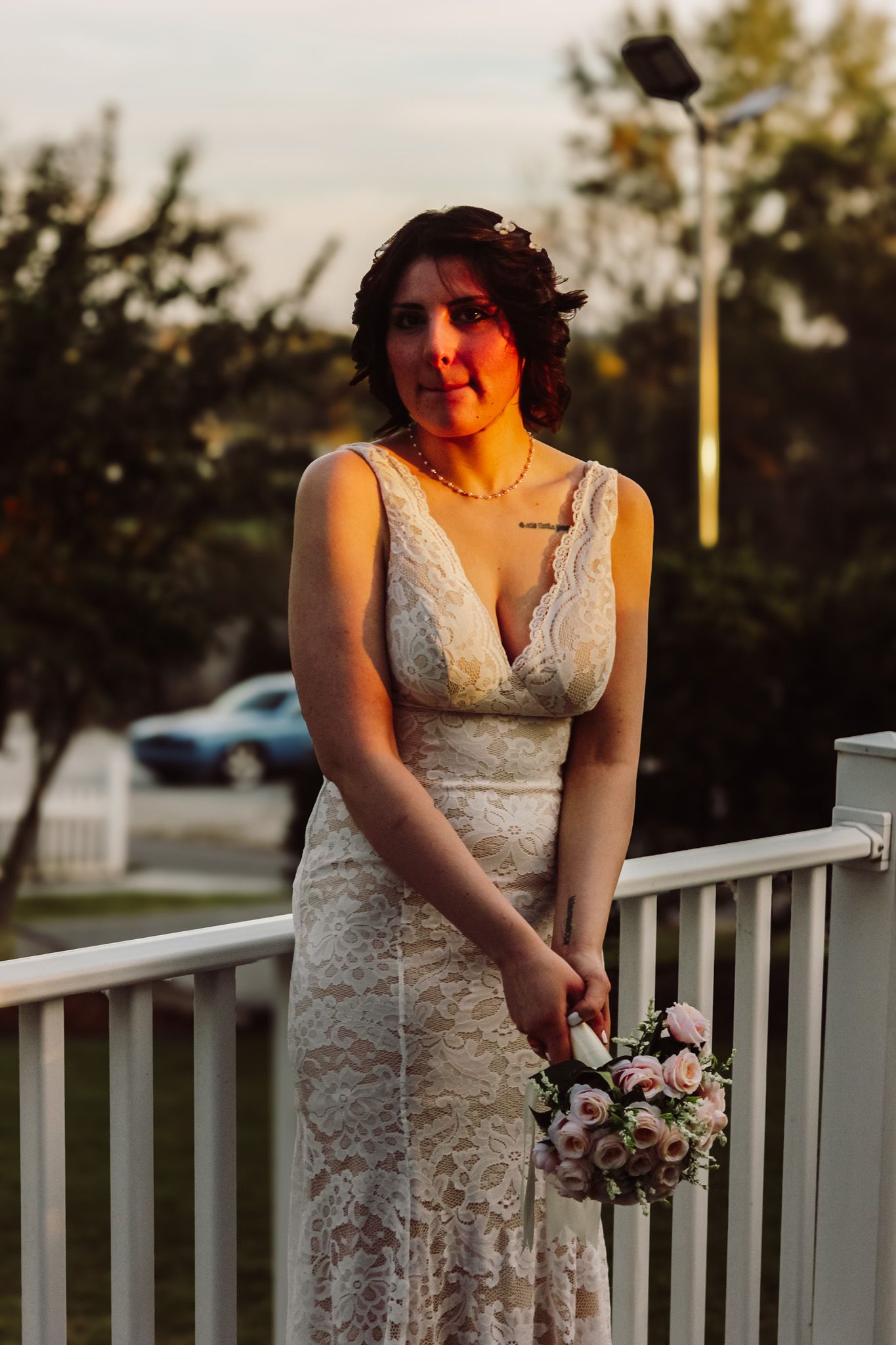 Bride in white lace dress holding bouquet at sunset on outdoor porch.