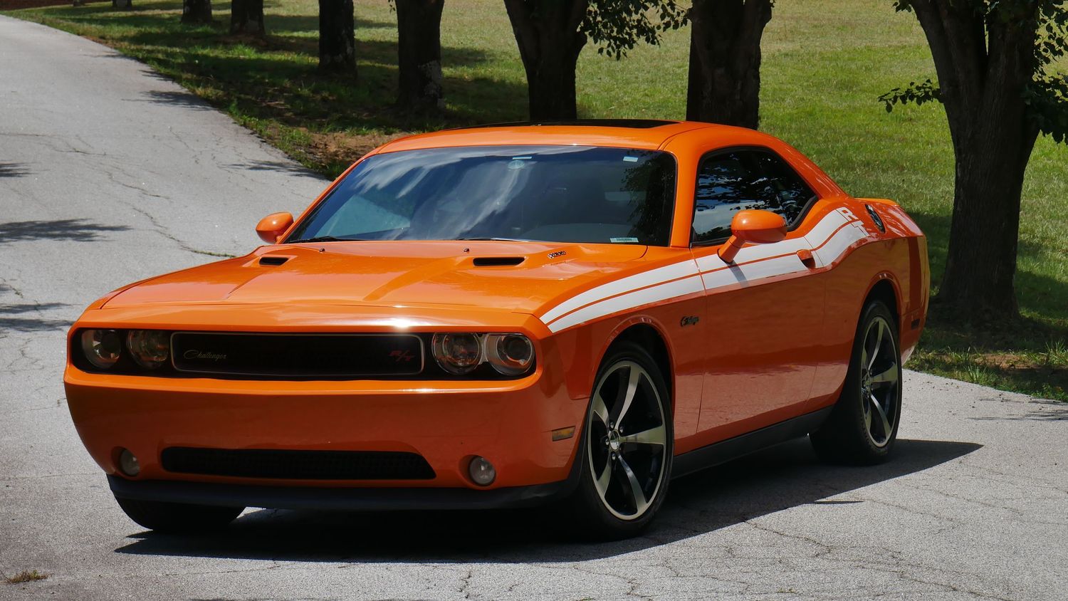 Orange Dodge Challenger with black wheels photographed on a scenic tree-lined road.