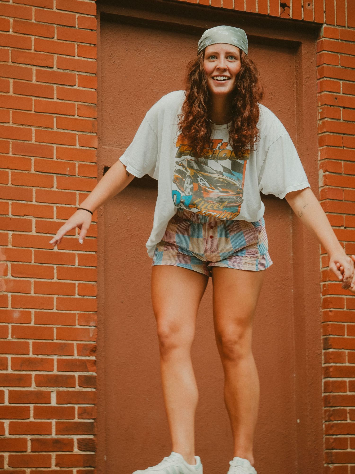 Person in casual summer outfit with gray beanie and tie-dye shirt poses playfully against brick wall in downtown setting.