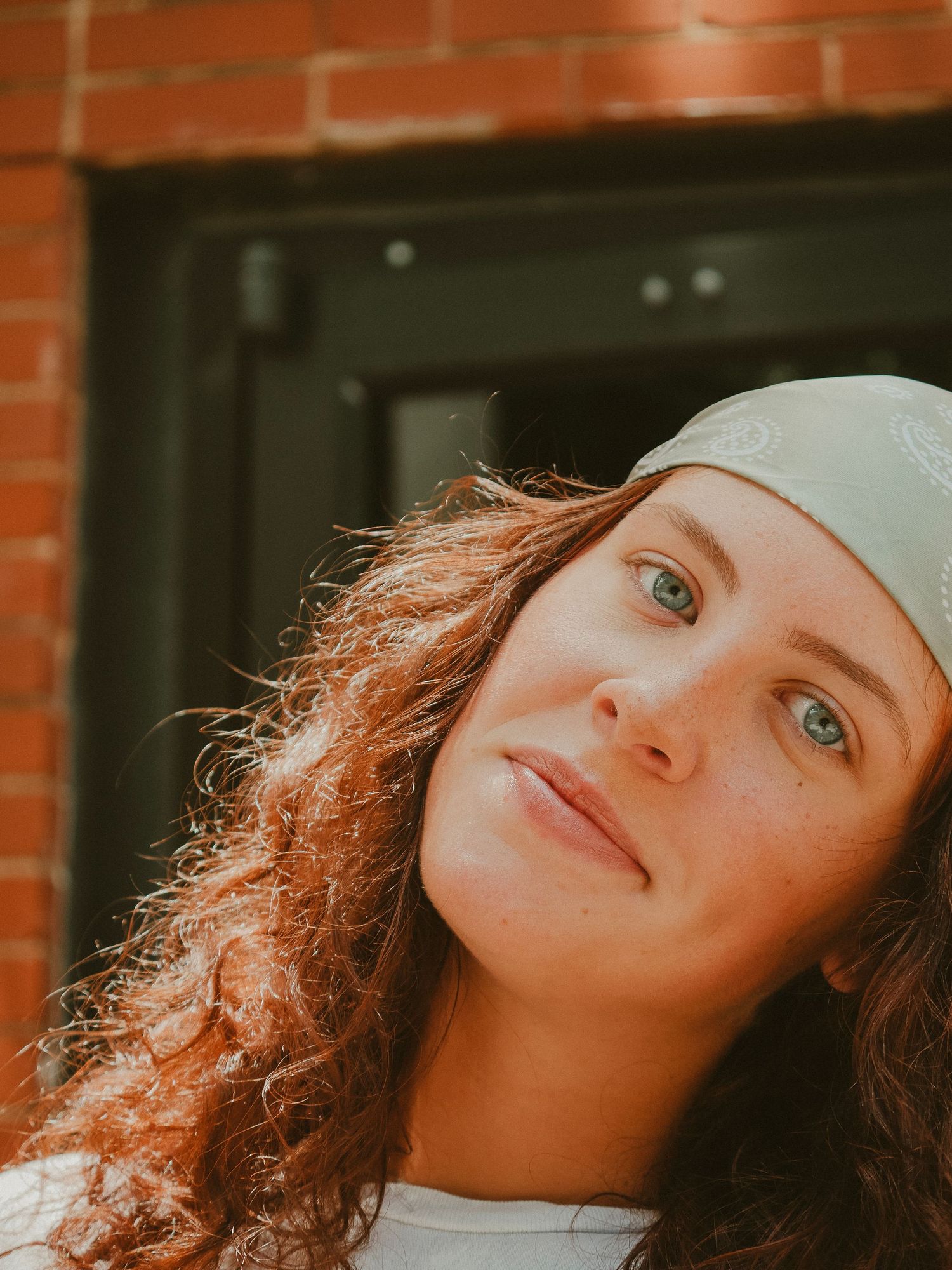 A portrait of someone with curly hair wearing a white beanie and white top against a brick wall in warm lighting.