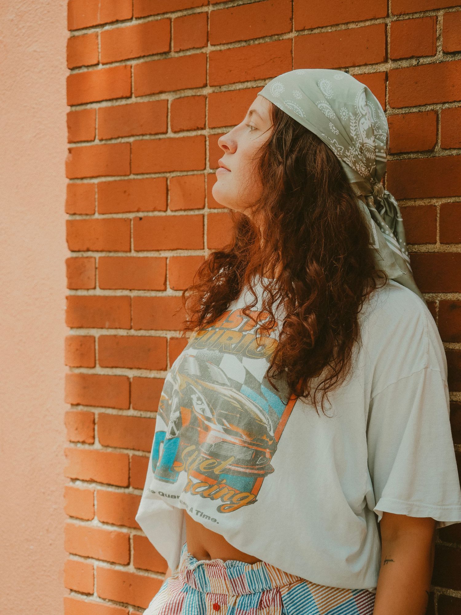 Person in a bandana and casual t-shirt leaning against a brick wall in a dreamy vintage-filtered outdoor portrait.