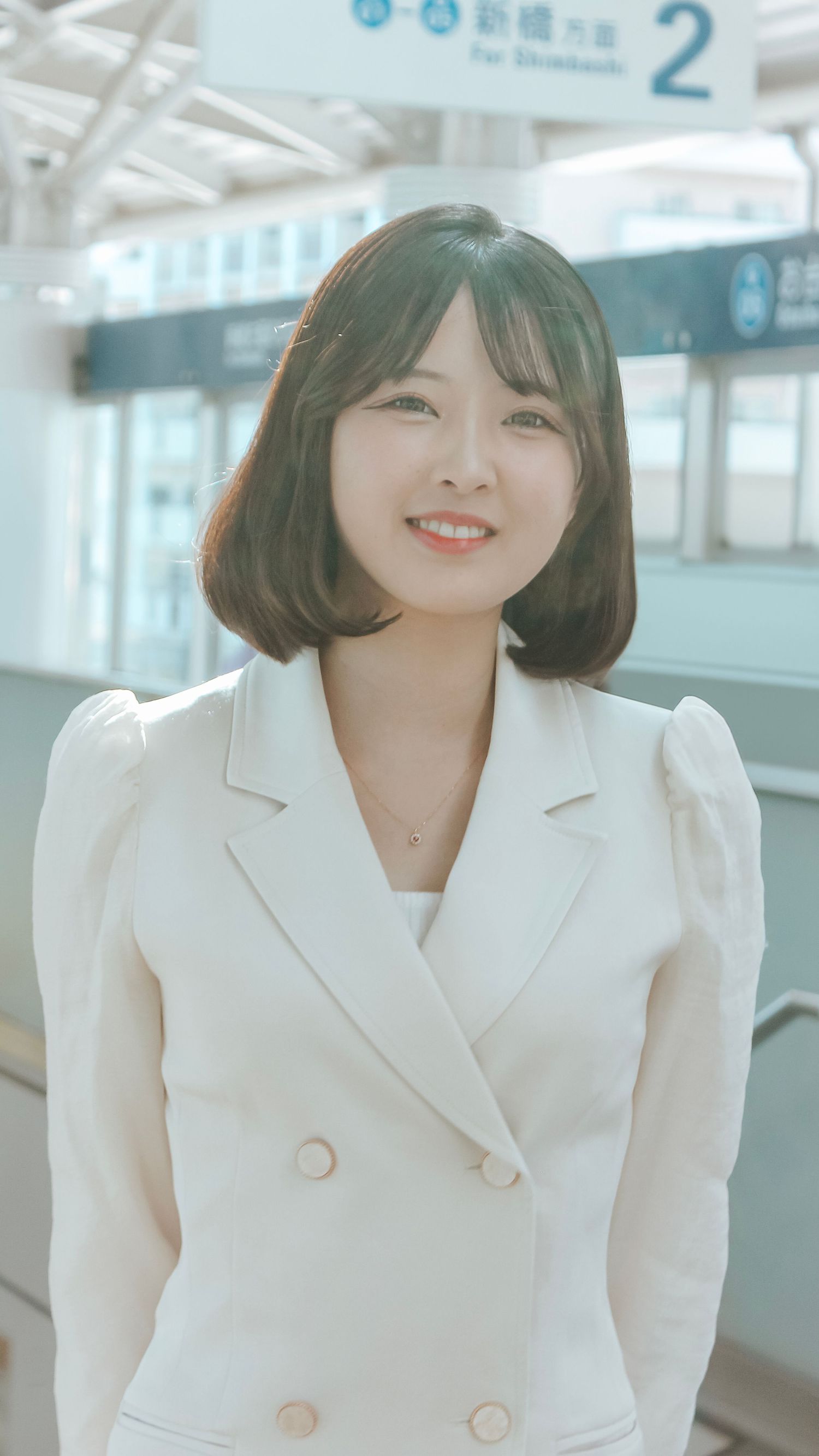 A person in a white blazer is standing in a bright modern corridor with a cheerful expression.