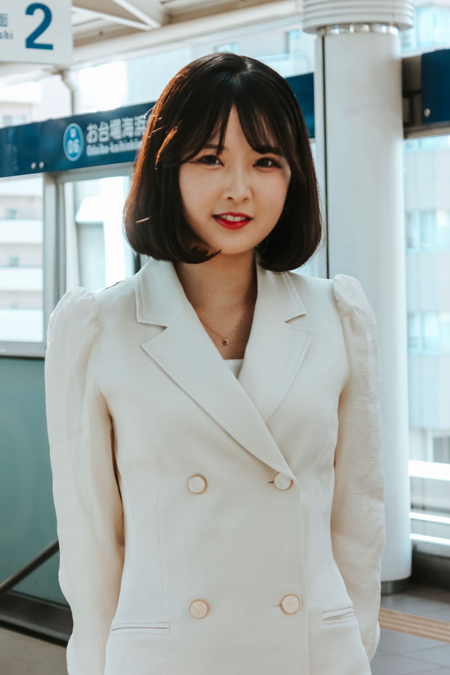 A person in a cream-colored double-breasted blazer stands in a subway station with Asian signage in the background.