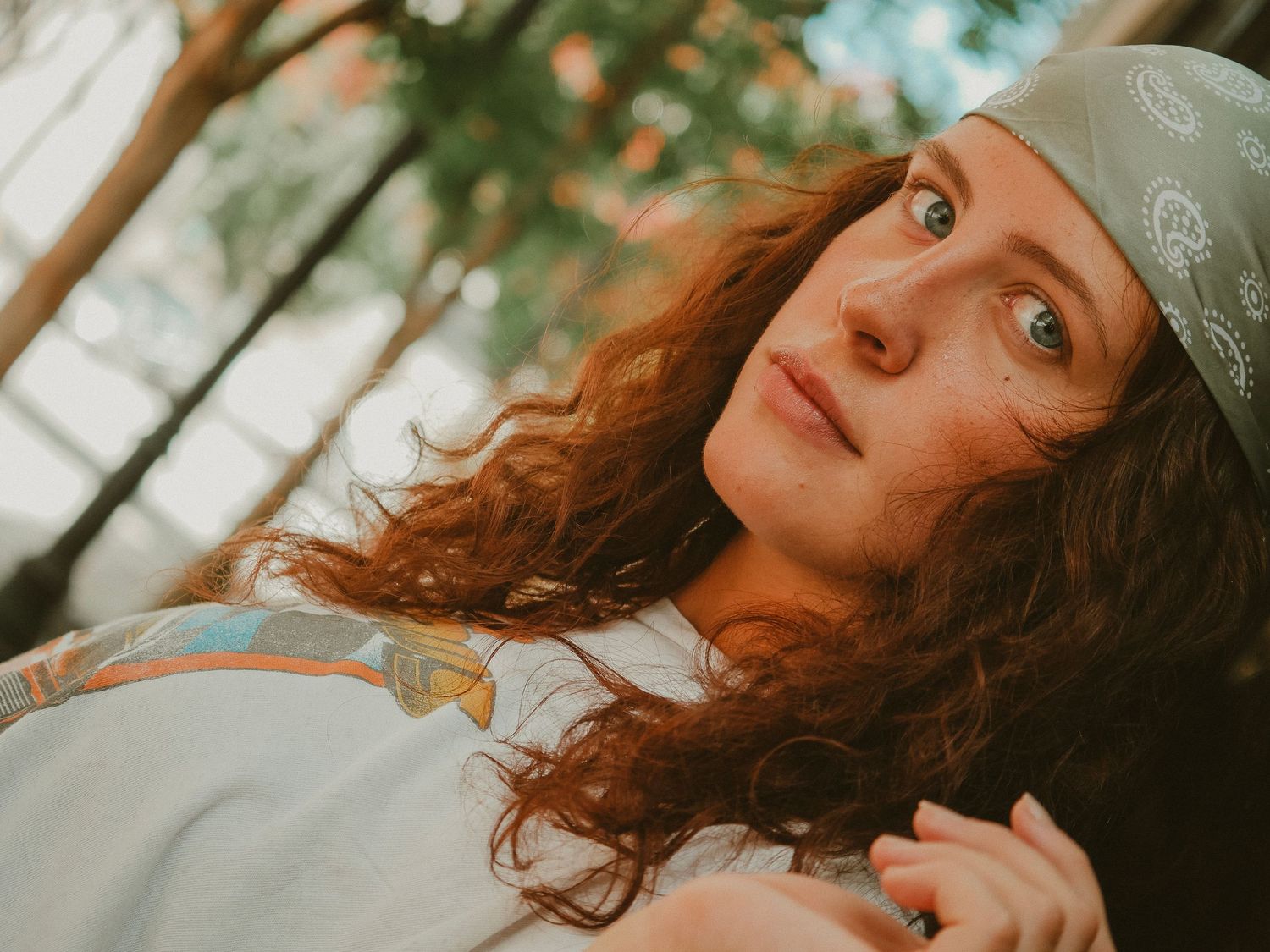 A person with curly auburn hair wearing a sage green beanie looks upward with a gentle smile in warm natural lighting.