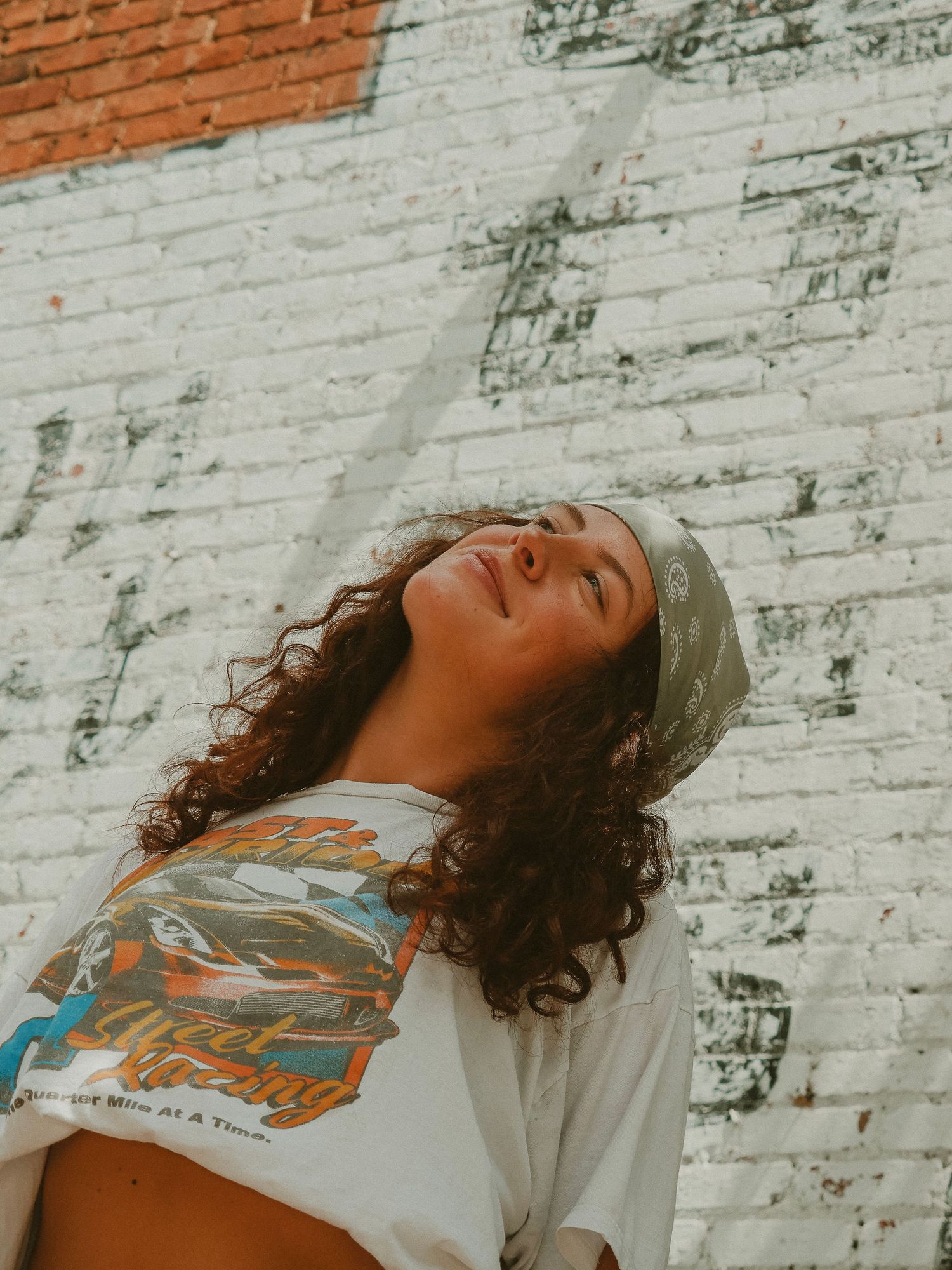 Person wearing a beanie and graphic t-shirt leans back against white painted brick wall in warm sunlight.