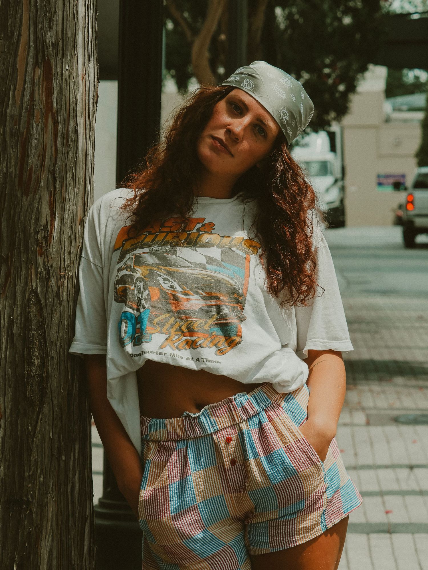 A person in a casual white t-shirt and patterned shorts leans against a wooden pole on an urban street.