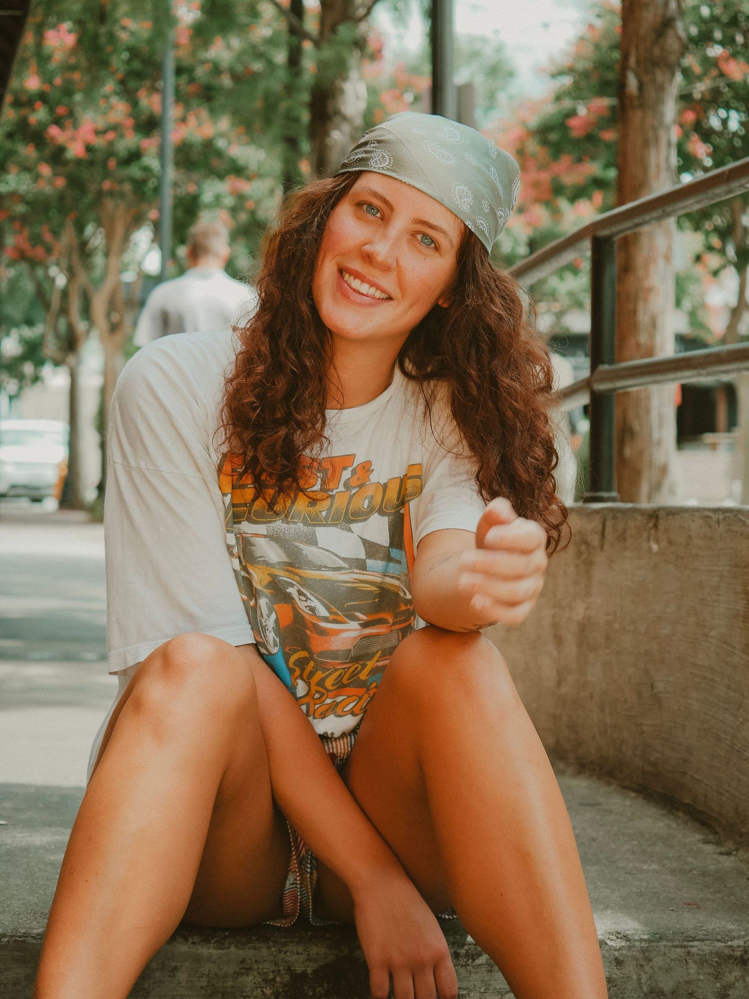 Someone in a beanie and vintage t-shirt sits casually outdoors with a warm smile in a sunlit urban setting.