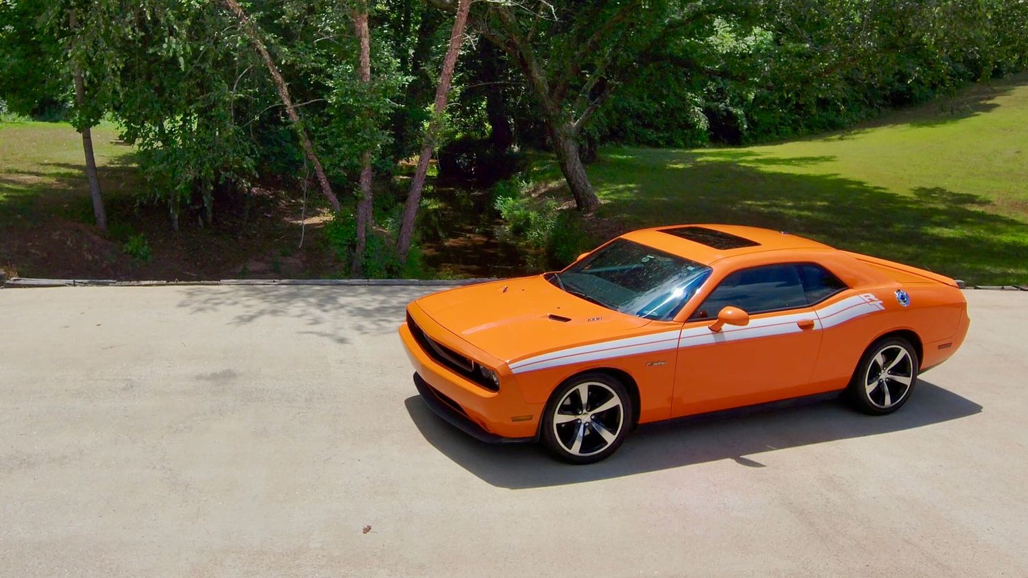 Modern orange muscle car parked on a curving asphalt driveway with trees and grass in the background.