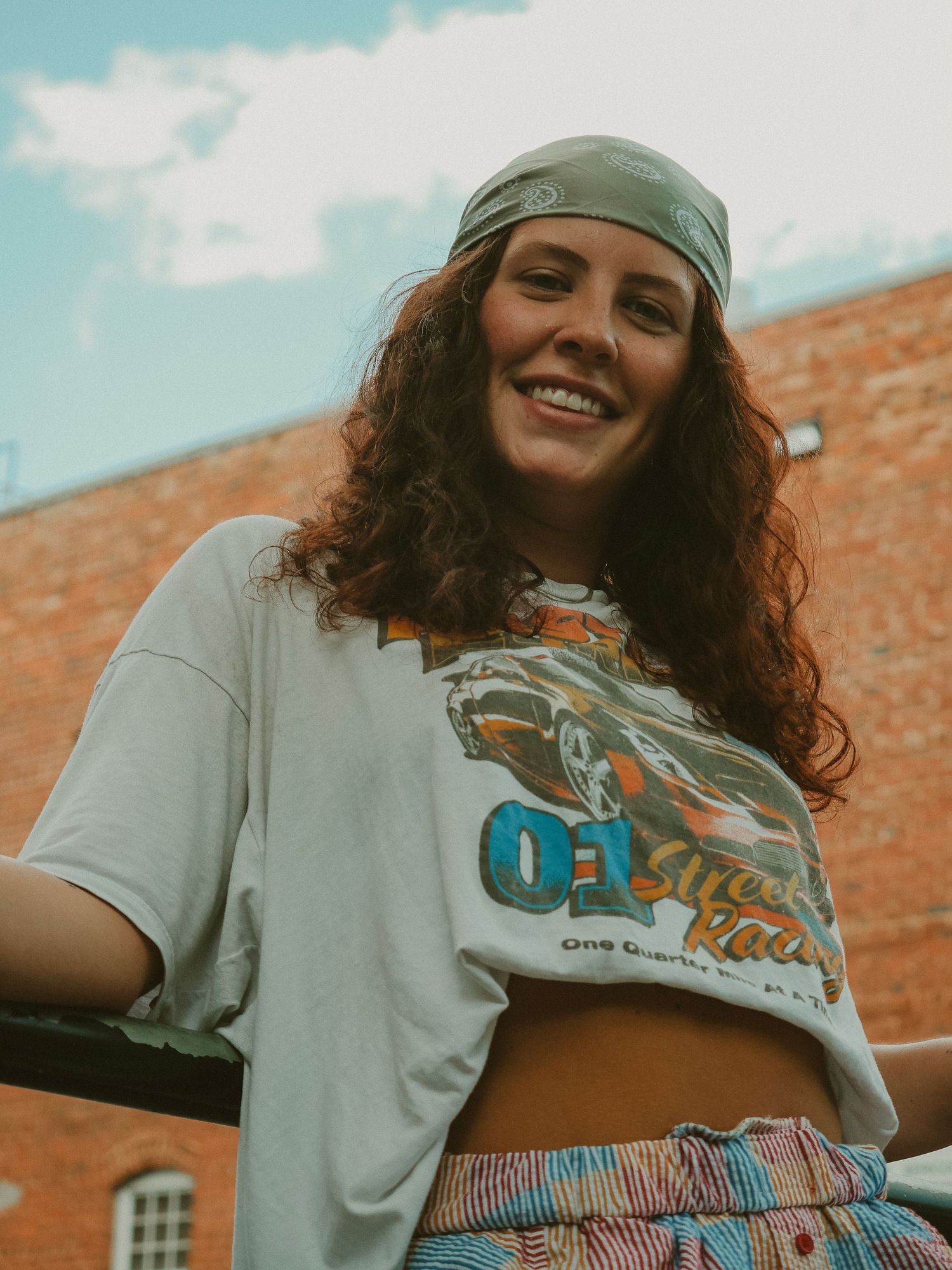 Woman in gray t-shirt and striped bottoms poses in front of brick wall on sunny day.