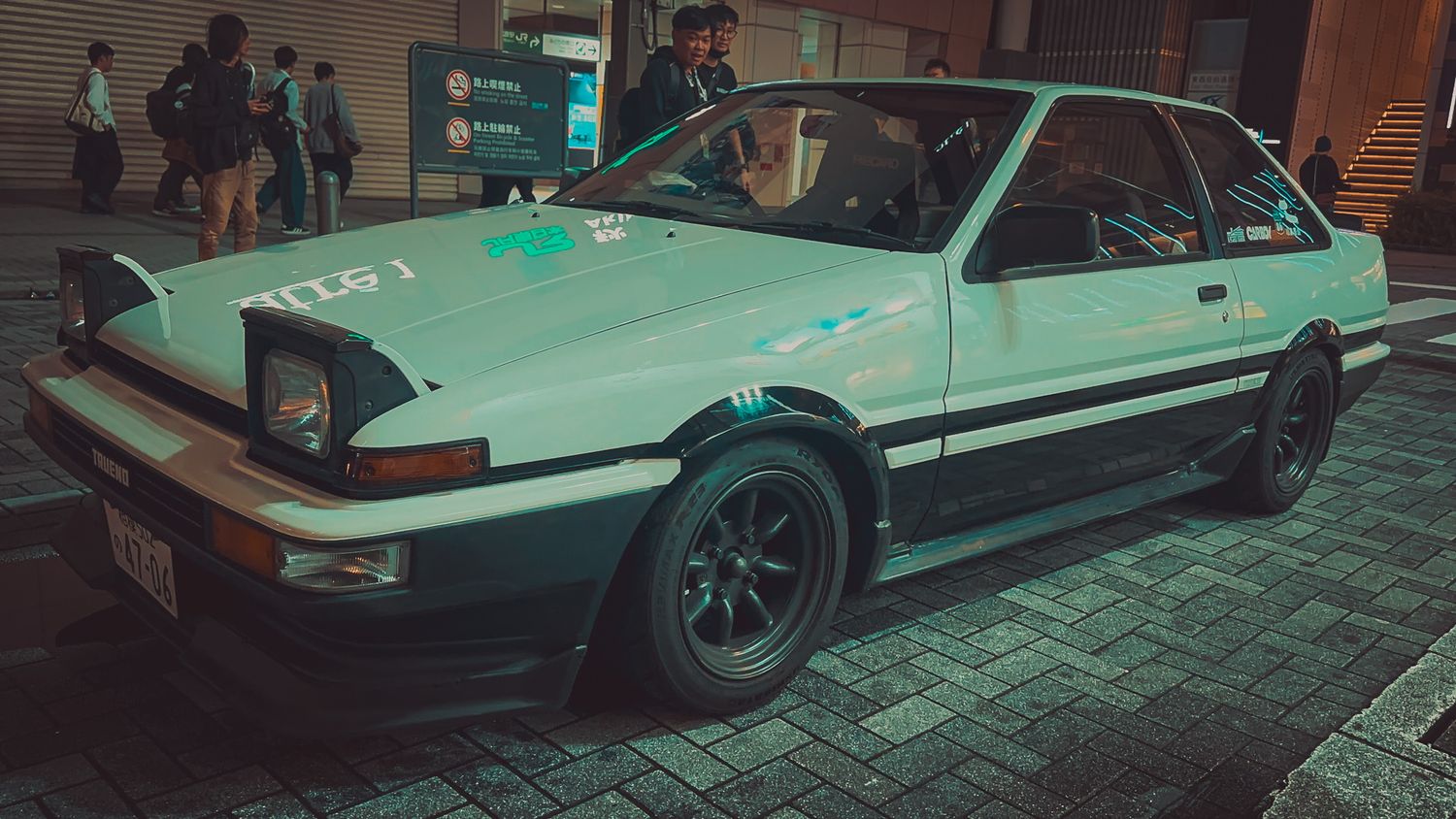 Row of modified Japanese sports cars displayed on cobblestone street at dusk.