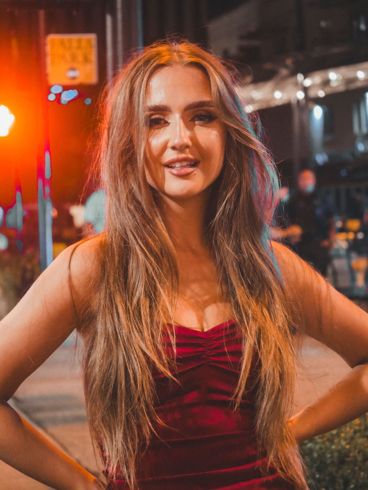 A young woman in a red dress smiles while standing on a city street at night with warm lighting and bokeh effects.