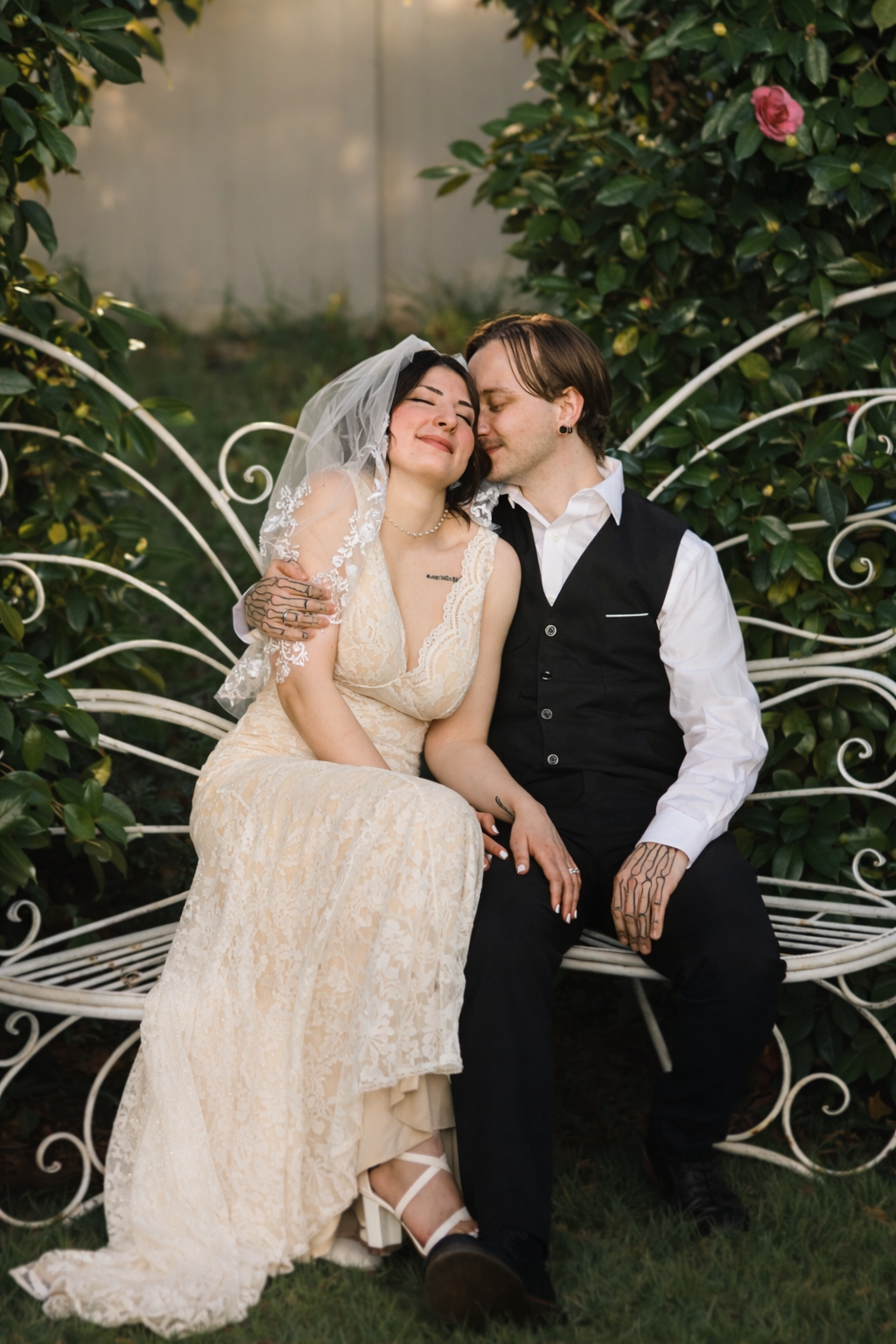 A romantic wedding couple shares intimate moments on a decorative white metal garden bench surrounded by greenery.