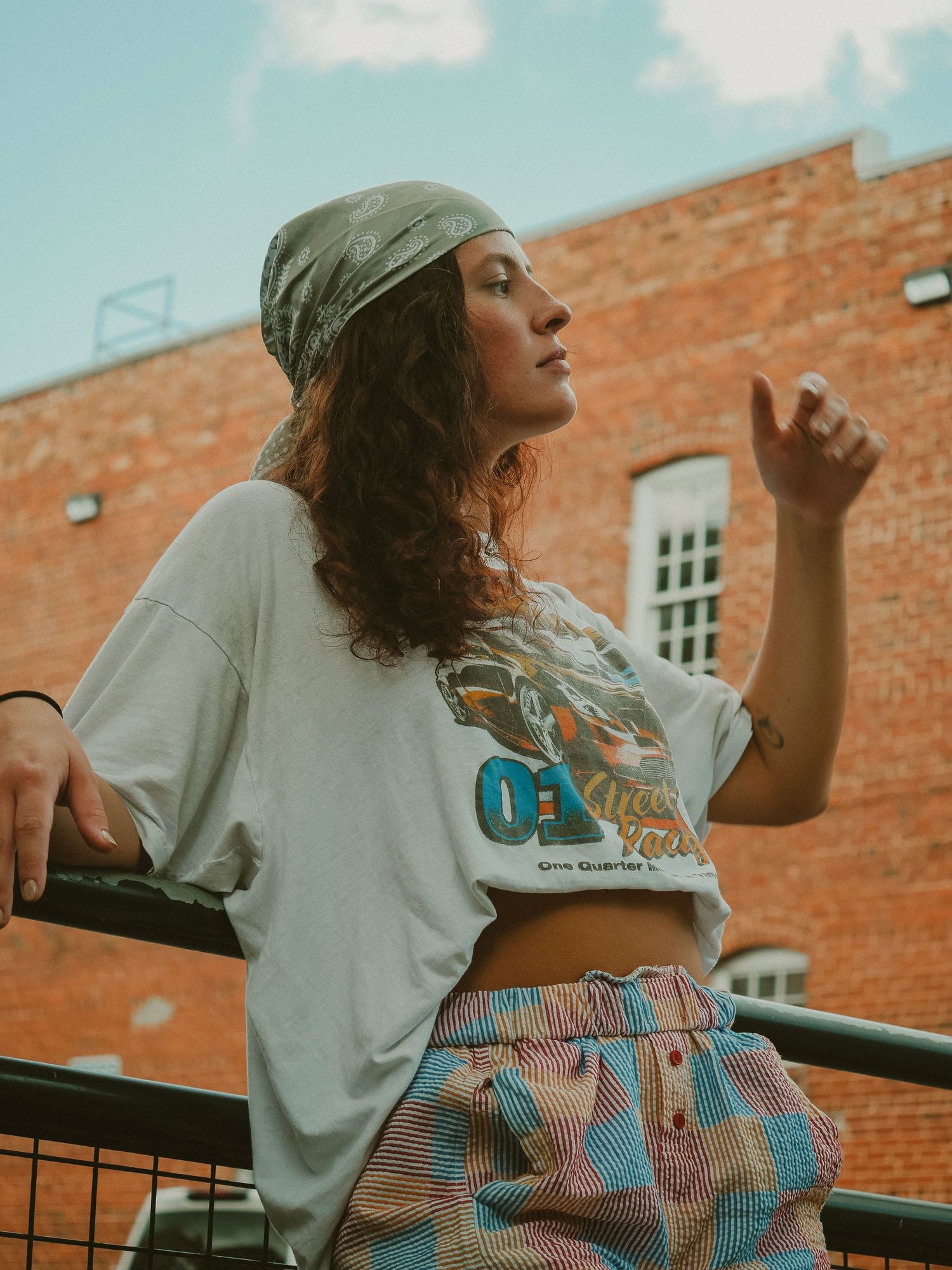 A person in a crop top and patterned shorts poses against a brick building backdrop on a sunny day.