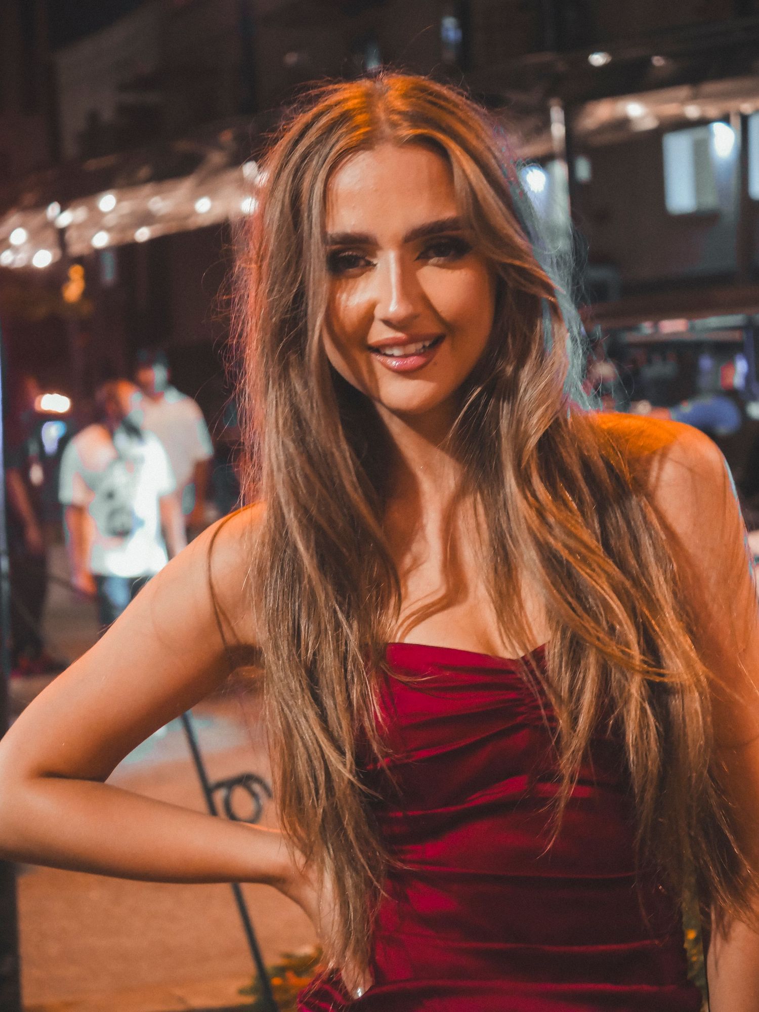 A woman in a red dress poses for a nighttime photo with string lights in the background.