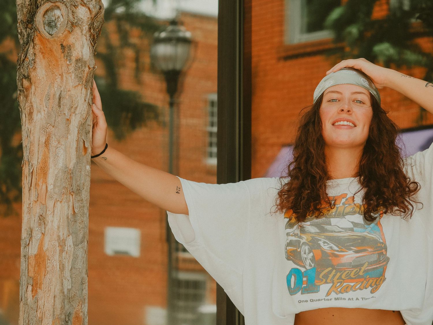Carefree woman with curly hair and white headband wearing retro t-shirt poses joyfully against orange brick wall.