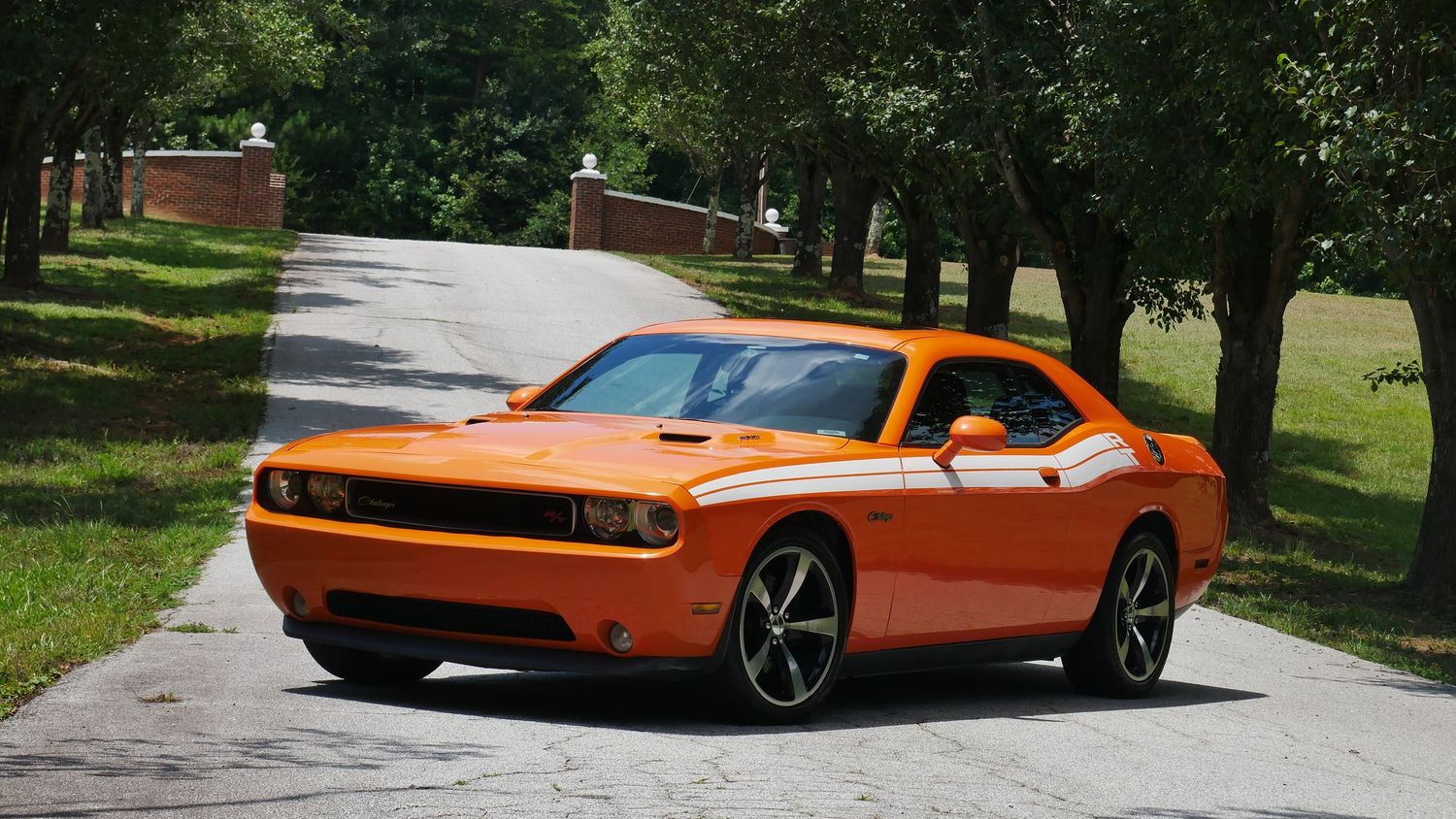 Orange Dodge Challenger muscle car parked on a tree-lined driveway in summer.