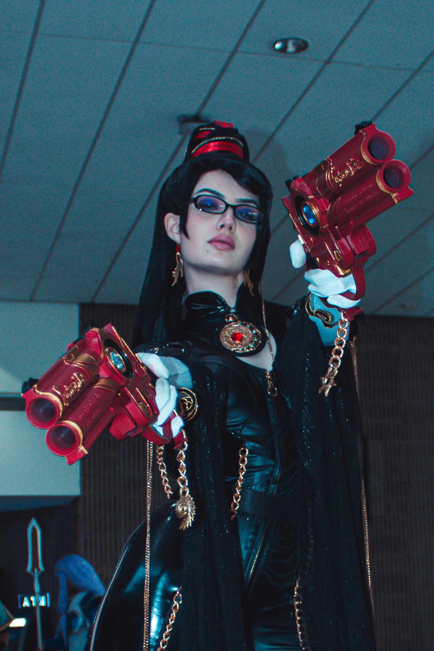 Cosplayer in black outfit wielding red prop guns in dramatic pose.