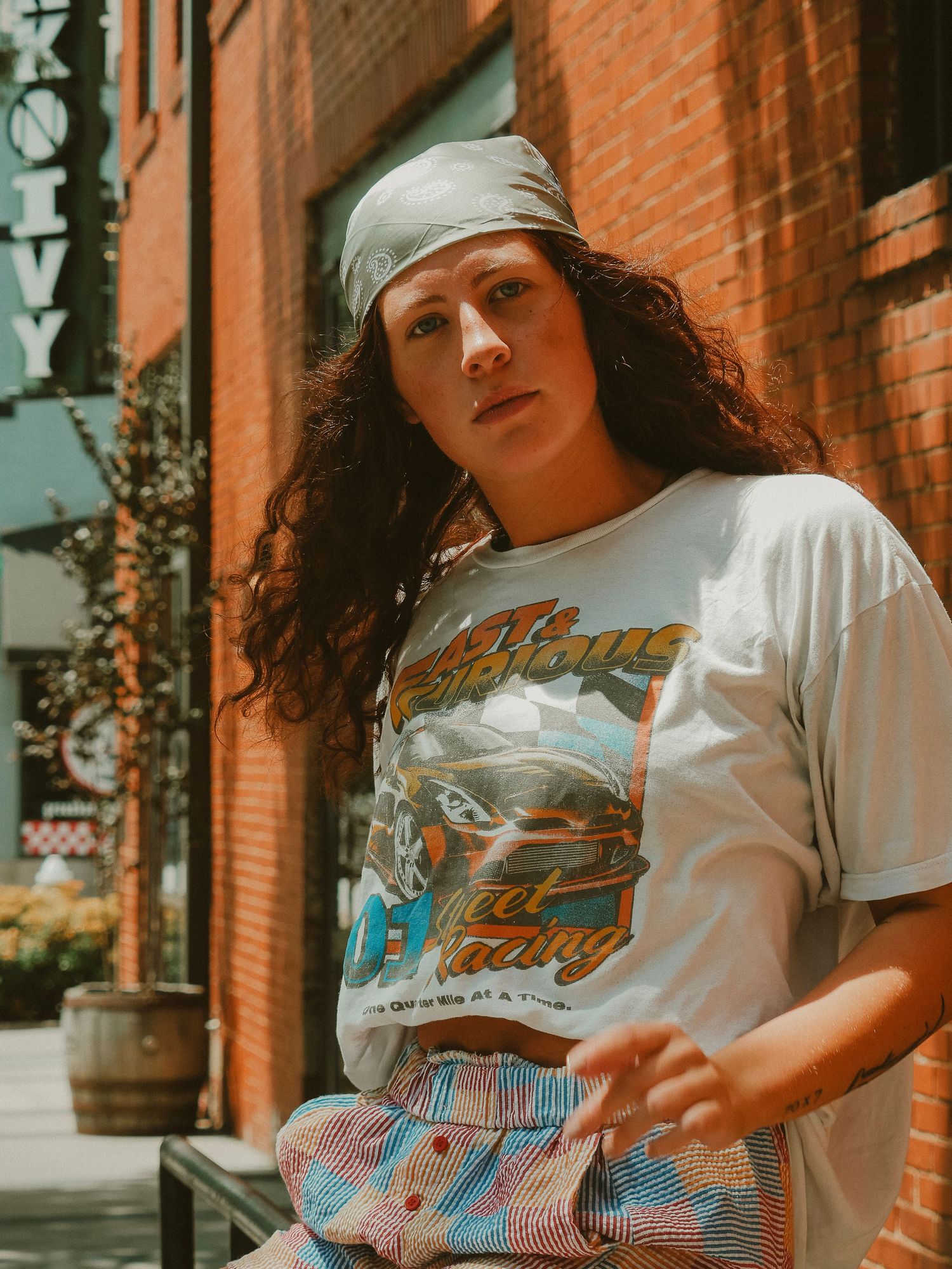 Woman in white graphic t-shirt and hat poses against rustic brick wall in urban setting.